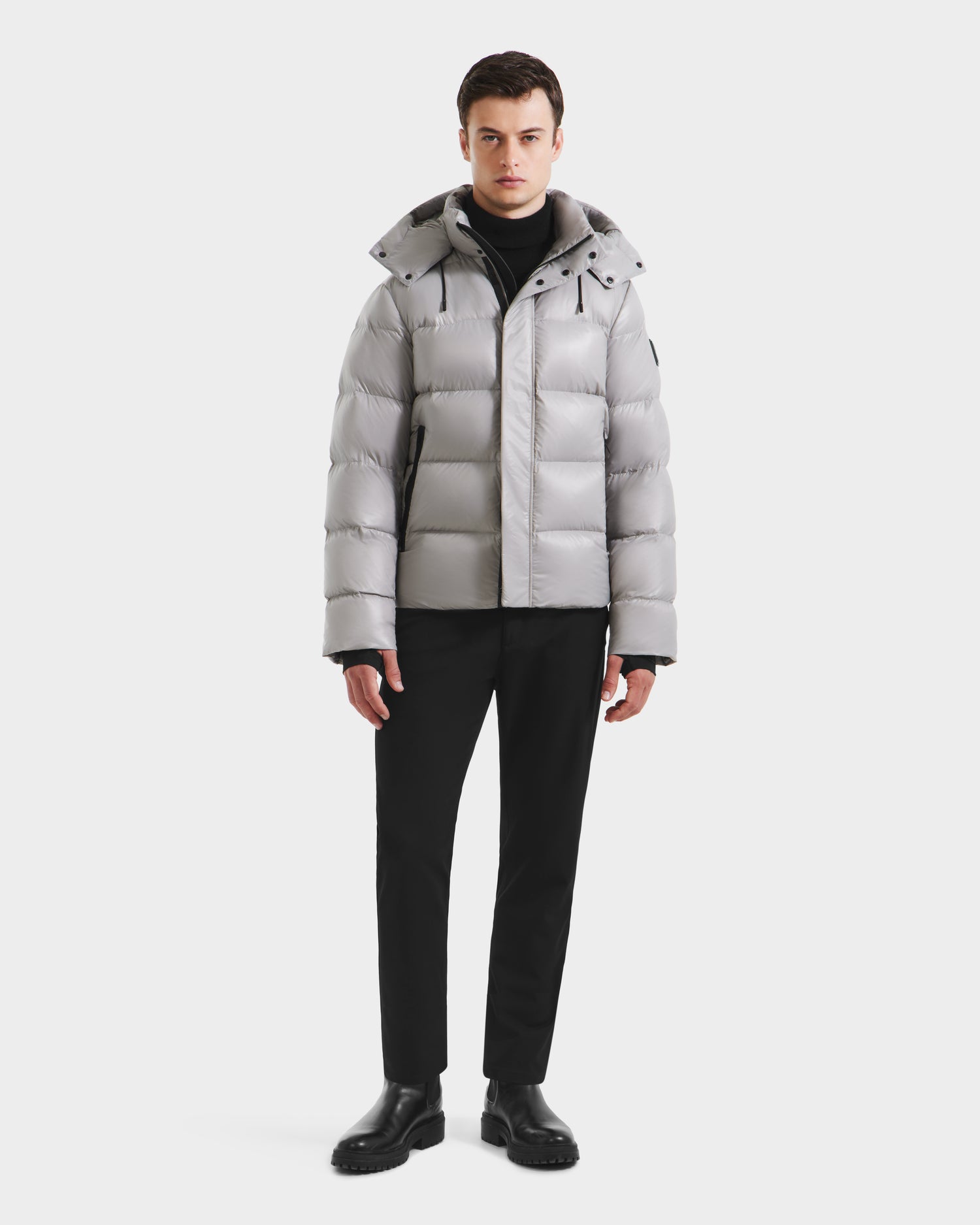 A man models the Rudsak JOEL S - 6124606 Light Grey down puffer jacket with a black turtleneck, pants, and boots against a white background, highlighting this stylish winter outerwear.