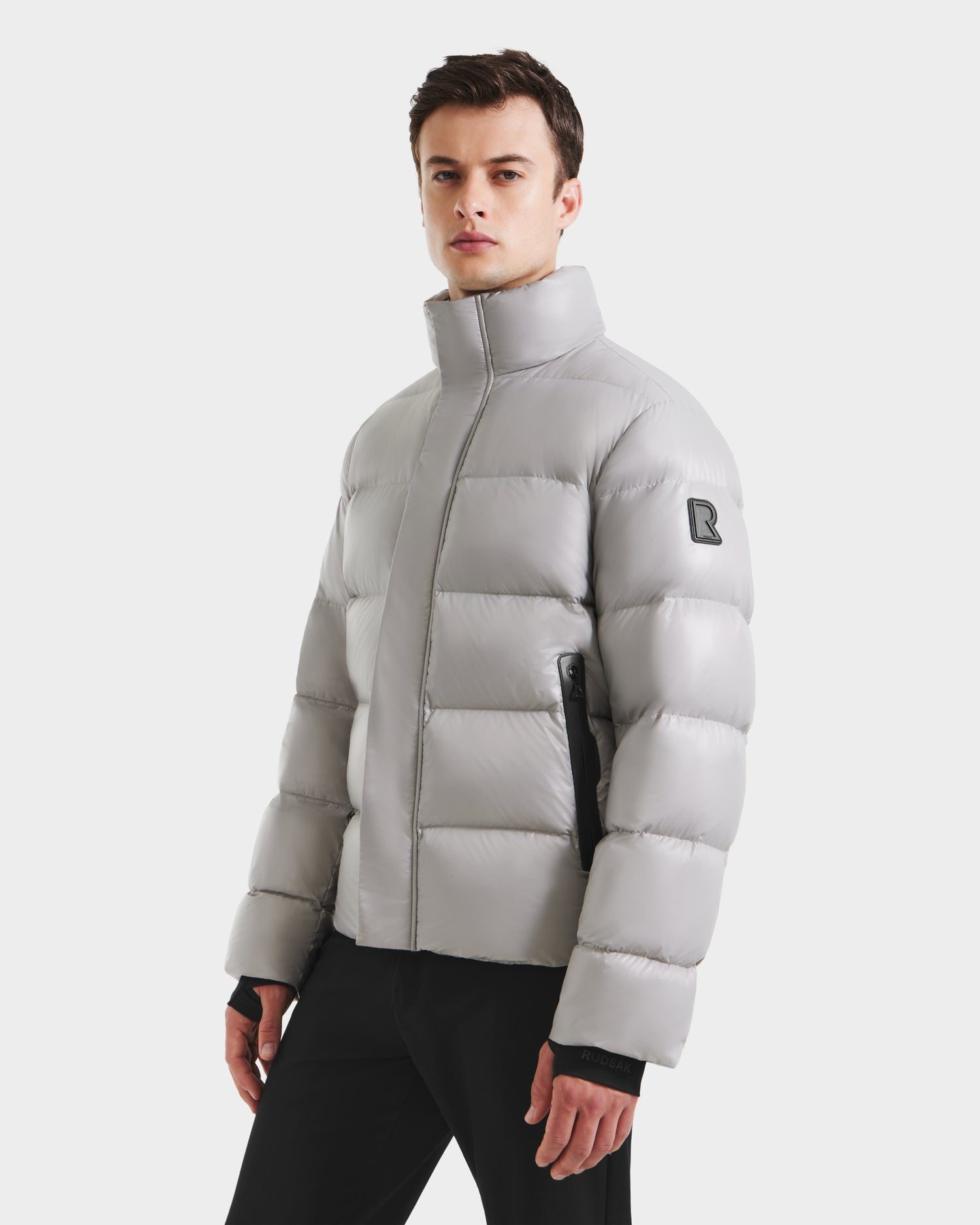 A man wearing the Rudsak JOEL S - 6124606 Light Grey premium down puffer jacket with a high collar and black zipper details, paired with black pants, stands against a plain white background.