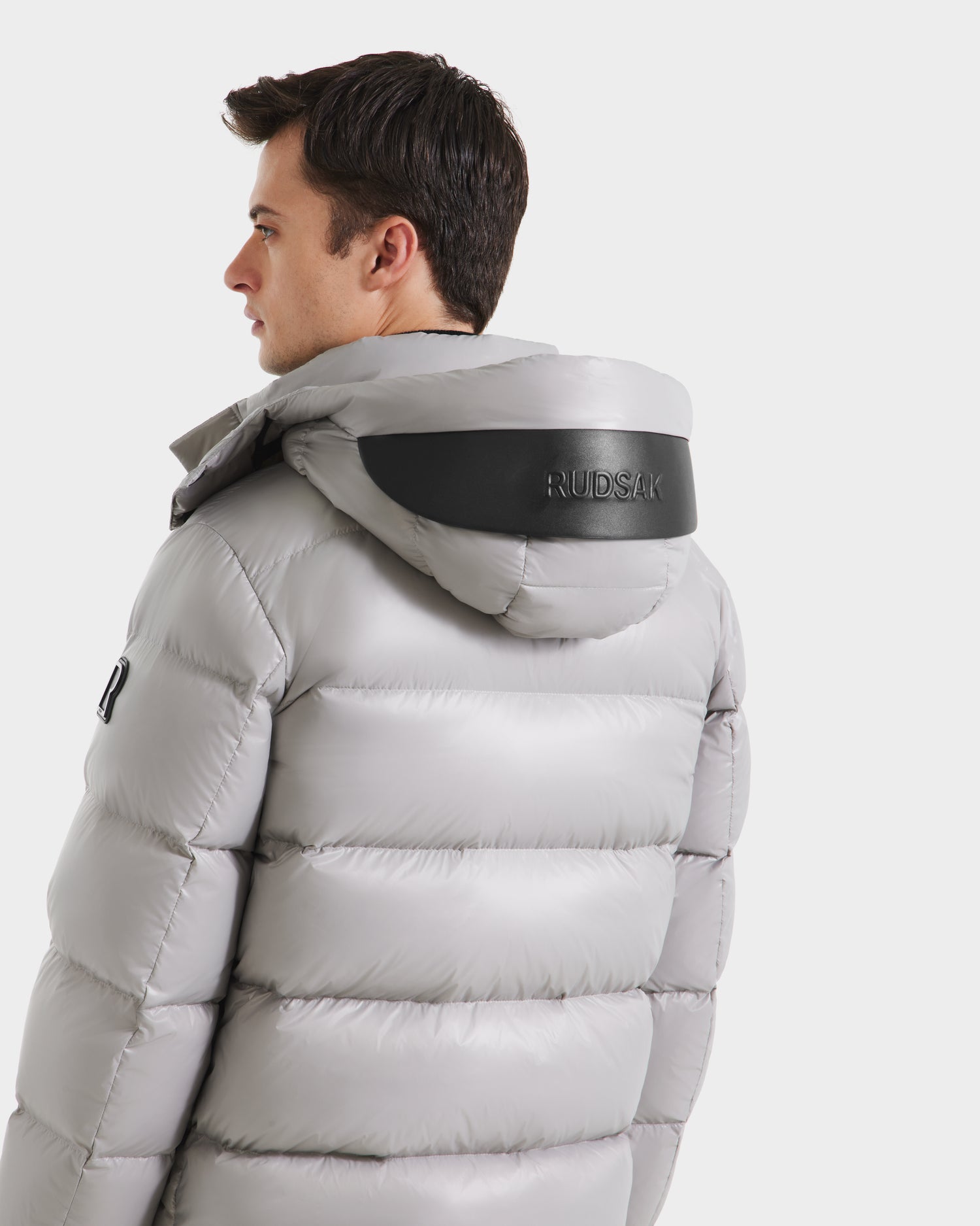 A man stands against a plain white background wearing the JOEL S - 6124606 Light Grey premium down puffer jacket by Rudsak, featuring a hood and a black Rudsak patch on the back—showcasing stylish winter outerwear.