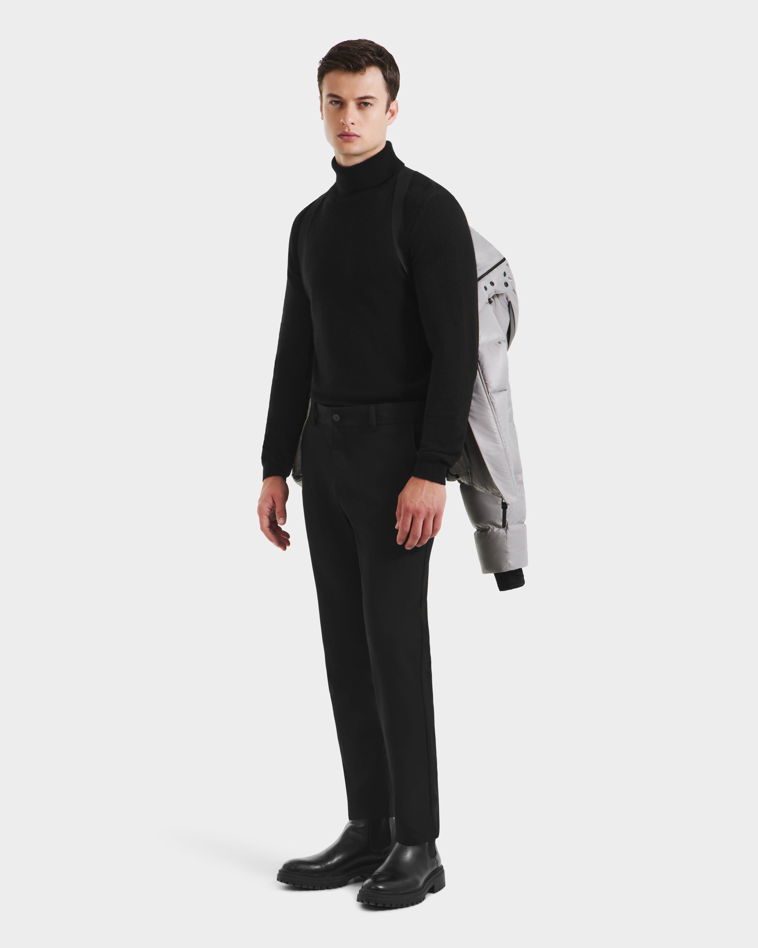 A man stands against a white background in black attire, holding the Rudsak JOEL S - 6124606 Light Grey premium down puffer jacket draped over his shoulder.