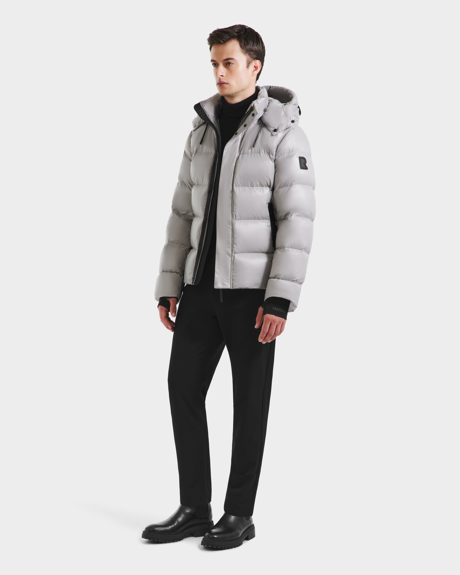 A young man models the Rudsak JOEL S - 6124606 light grey premium down puffer jacket with a hood, paired with black pants and boots against a plain white background, highlighting stylish winter outerwear.