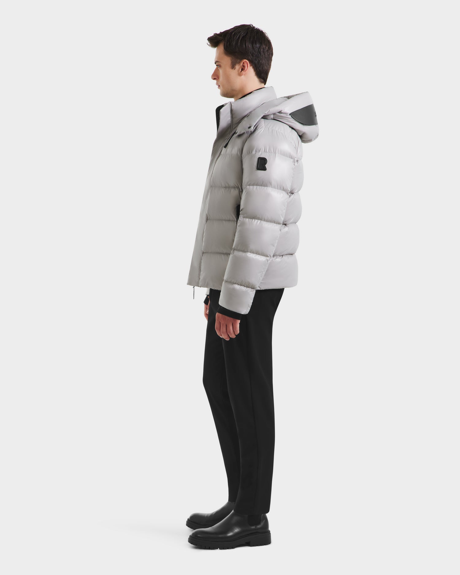 A man stands in profile against a white background, showcasing the Rudsak JOEL S - 6124606 Light Grey premium down puffer jacket with a hood, paired with black pants and boots—an example of stylish winter outerwear.