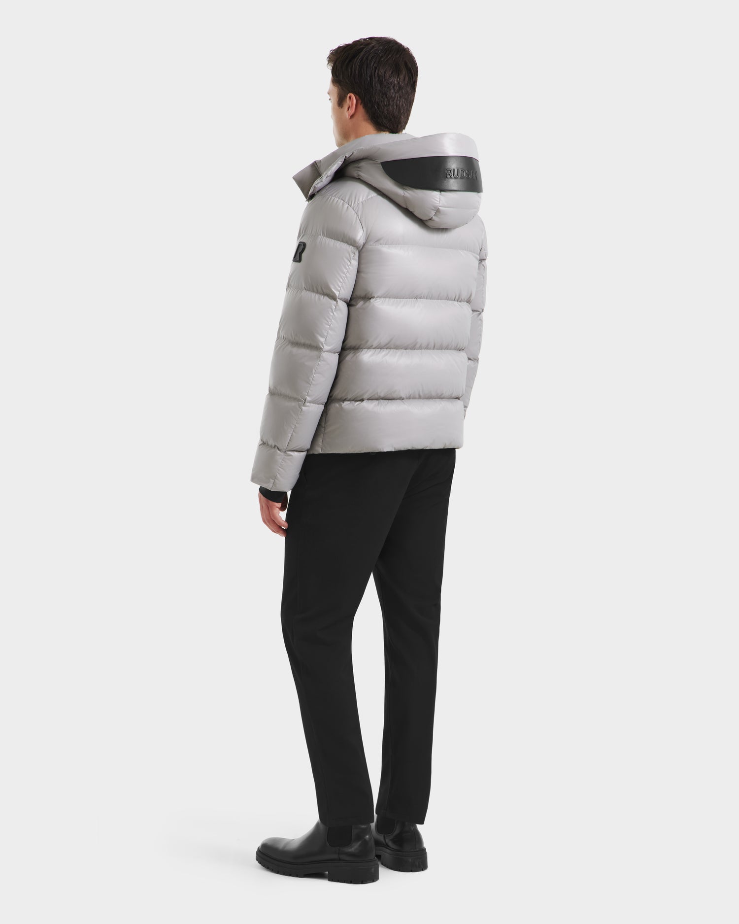 A person stands facing away, wearing the Rudsak JOEL S - 6124606 Light Grey premium down puffer jacket with a hood, paired with black pants and shoes against a plain white background.
