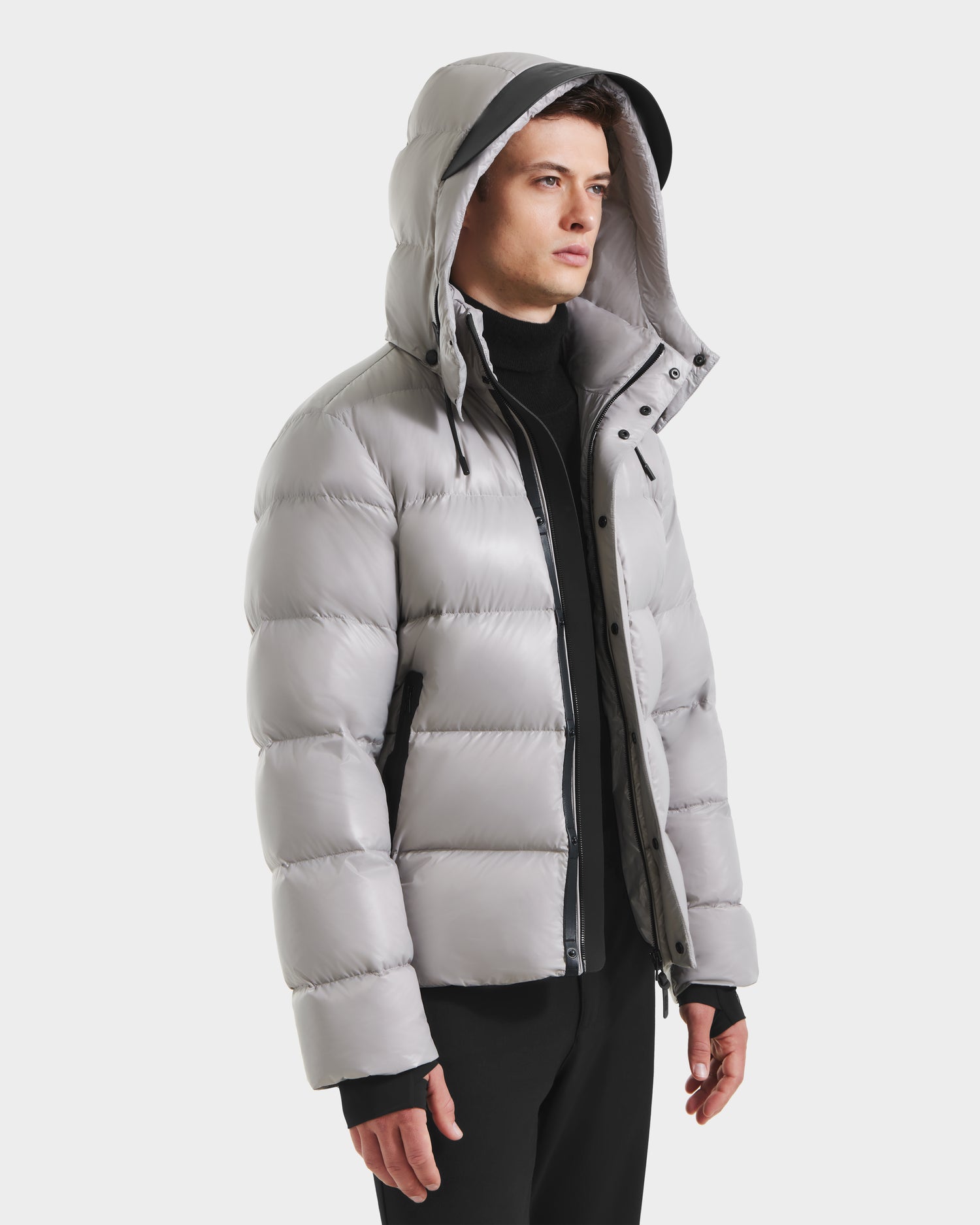 A person in the Rudsak JOEL S - 6124606 Light Grey puffer jacket with removable hood, paired with black pants and a black turtleneck, stands sideways against a white background.