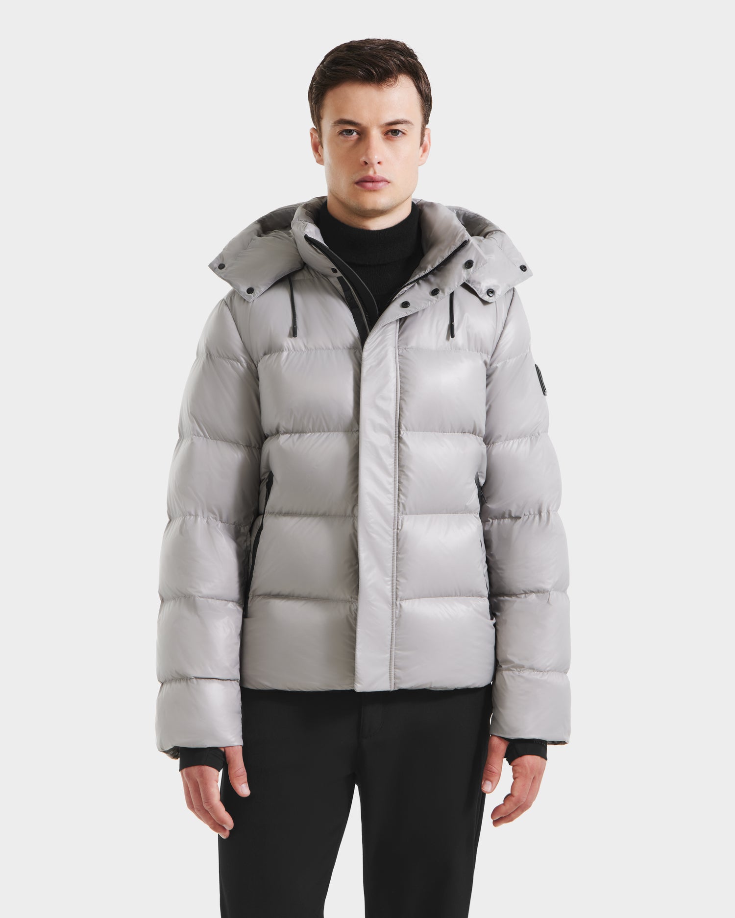 A young man faces forward in the Rudsak JOEL S - 6124606 Light Grey down puffer jacket with a hood and black pants, showcasing stylish winter outerwear against a plain white background.