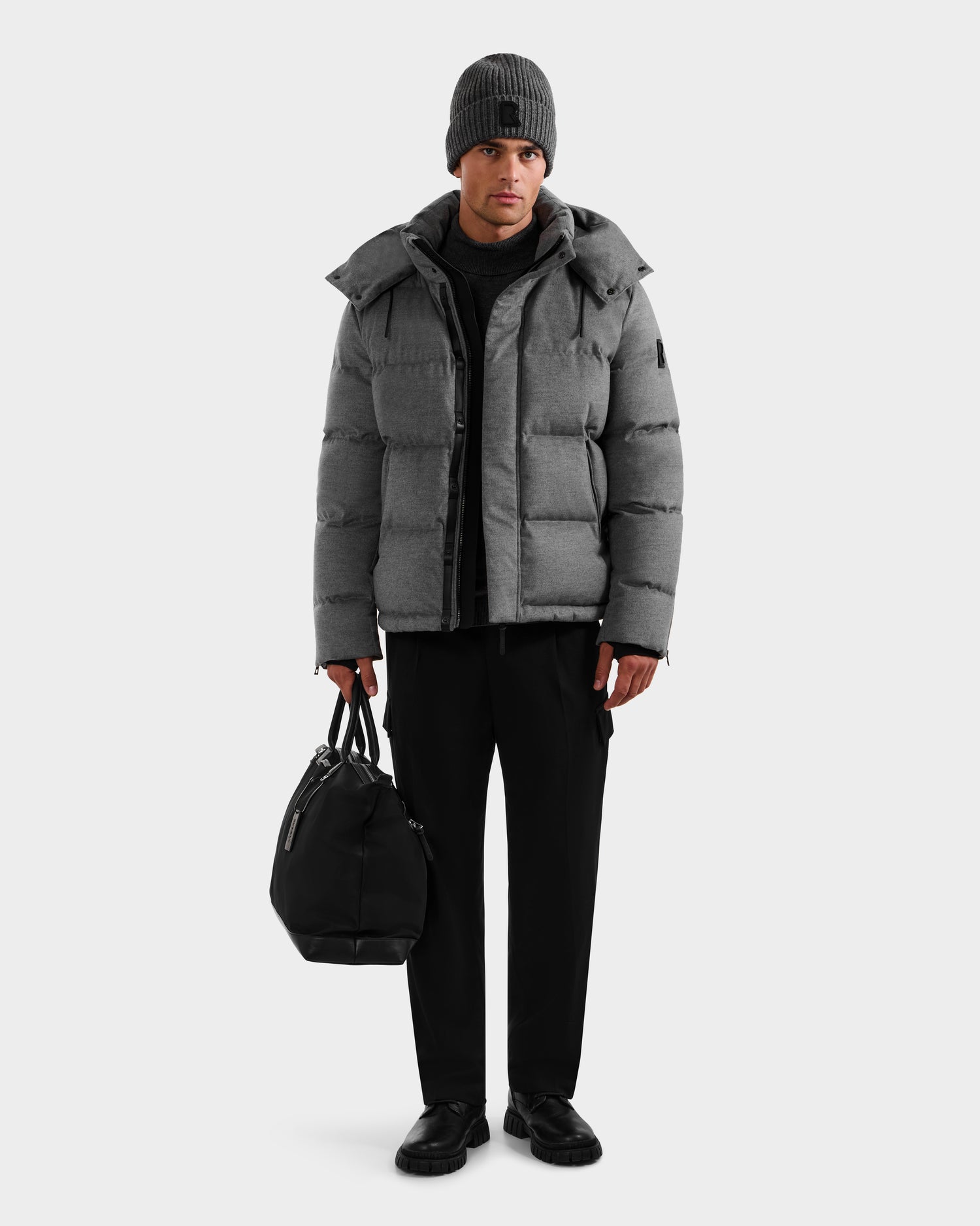 A man stands facing forward against a white background, wearing the Rudsak JOEL W - 6125683 Heather Charcoal down jacket with a removable hood, paired with black pants, black shoes, a gray beanie, and holding a black duffel bag in his left hand.