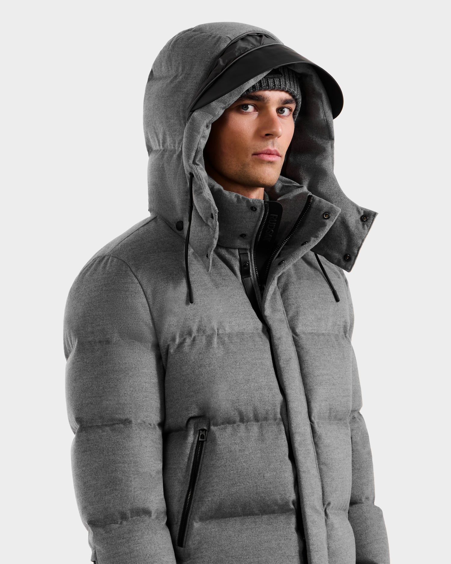 A man wears the Rudsak JOEL W - 6125683 Heather Charcoal wool-blend down puffer jacket with a removable hood and a black beanie, looking slightly to the side against a plain light background.