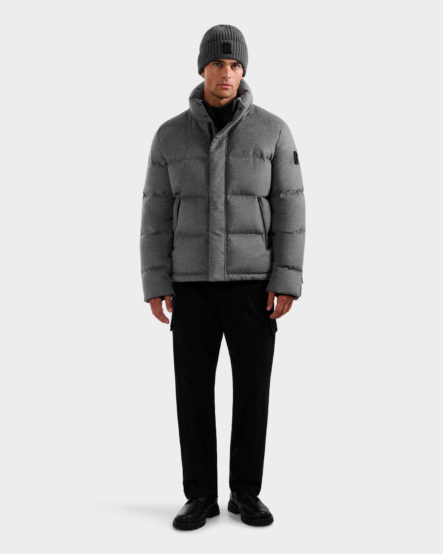 A man stands facing forward against a plain light background, wearing the Rudsak JOEL W - 6125683 Heather Charcoal wool-blend down puffer with removable hood, black pants, black shoes, and a gray knit beanie.