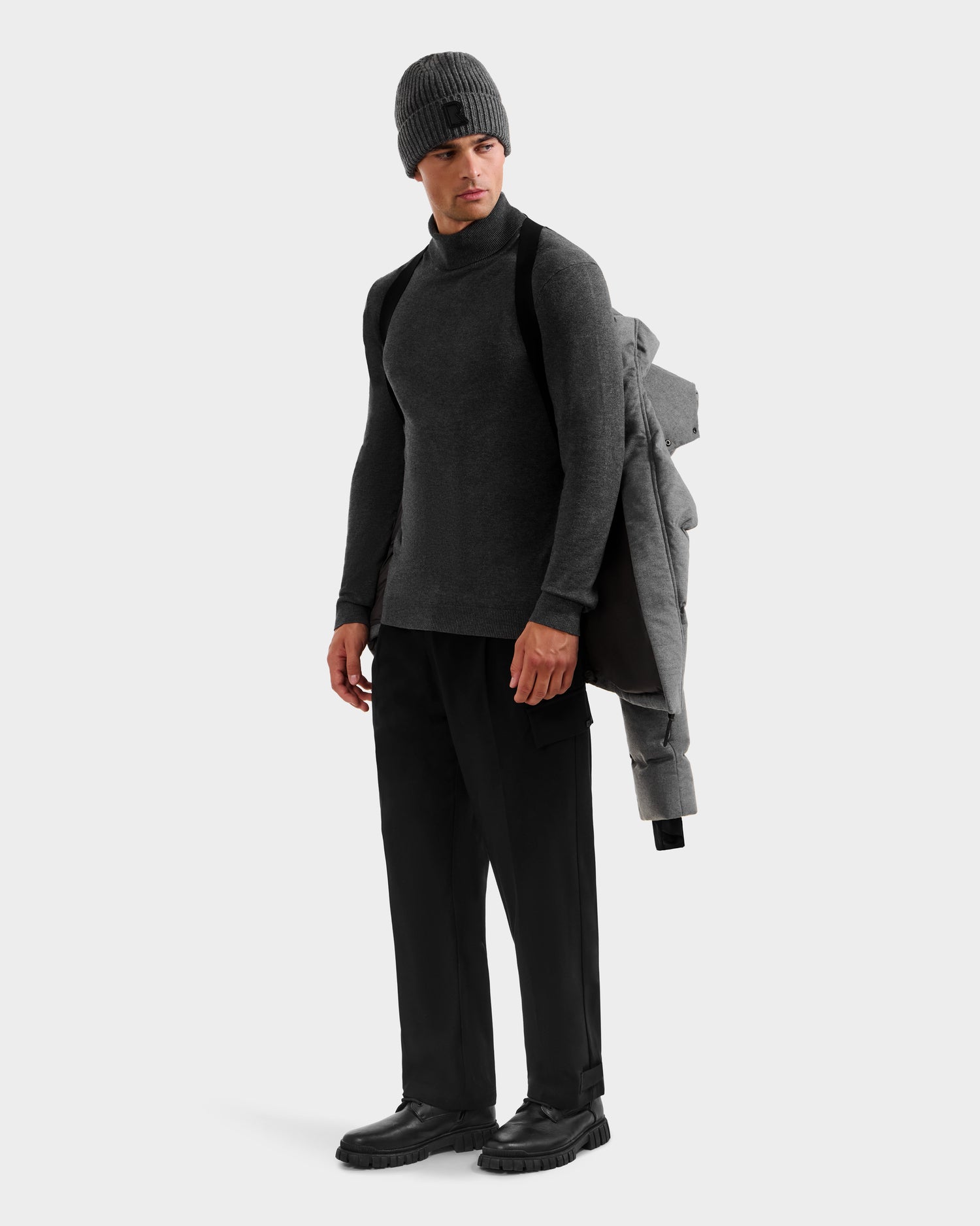 A man stands against a plain light background, holding the Rudsak JOEL W - 6125683 Heather Charcoal men’s wool-blend down puffer with a removable hood over his shoulder, wearing a gray knit beanie, dark turtleneck, black pants, and black shoes.