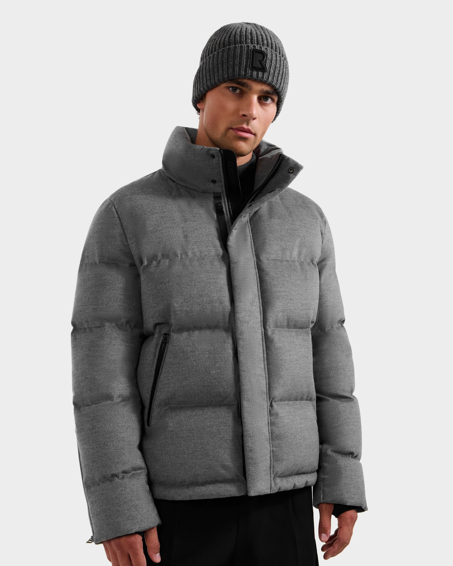 A man wearing the Rudsak JOEL W - 6125683 Heather Charcoal puffer and a gray knit "R" beanie stands against a plain light background, looking directly at the camera with a neutral expression.