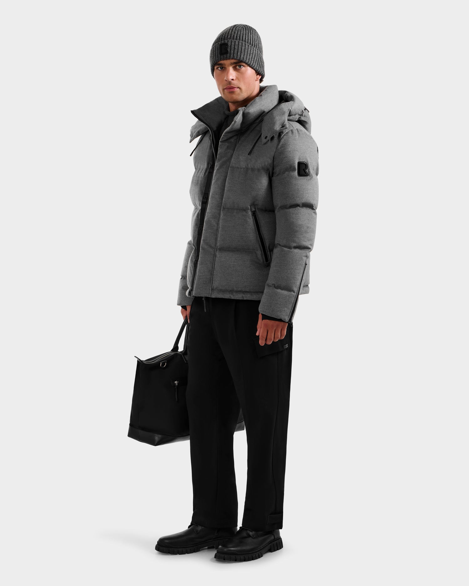 A man stands against a white background wearing the Rudsak JOEL W - 6125683 Heather Charcoal wool-blend down puffer with removable hood, black pants and shoes, a gray knit beanie, and holding a black bag by his side.