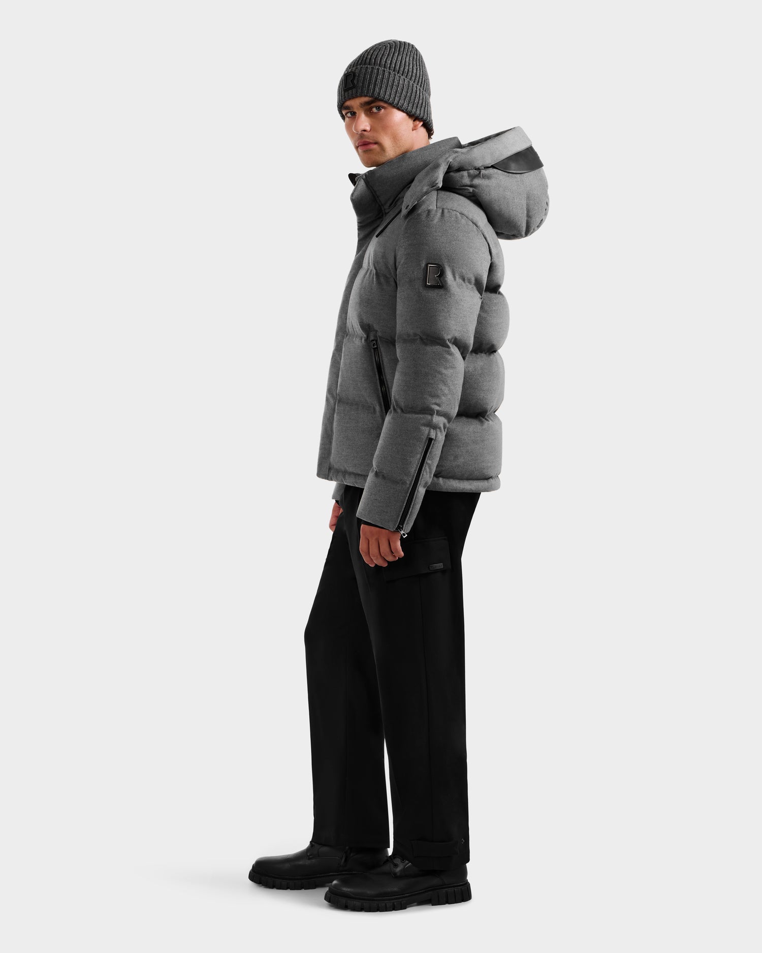 A person wearing the Rudsak JOEL W - 6125683 Heather Charcoal men’s puffer with a removable hood, black pants, black boots, and a gray knit beanie stands looking to the side against a plain white background.