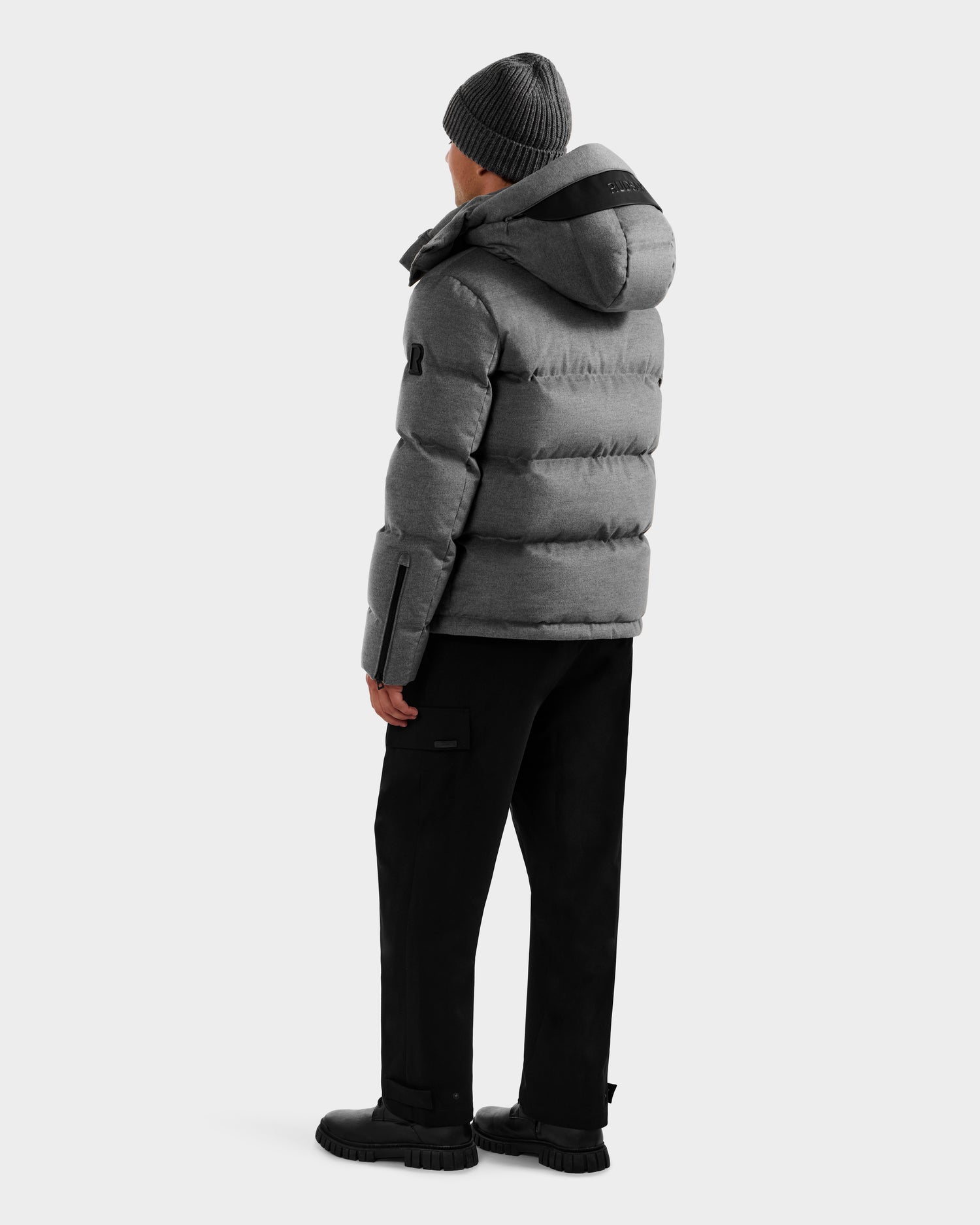 Wearing the Rudsak JOEL W - 6125683 Heather Charcoal wool-blend down puffer, black pants, black shoes, and a gray knit beanie, a person stands facing away and looks left against a plain light background.