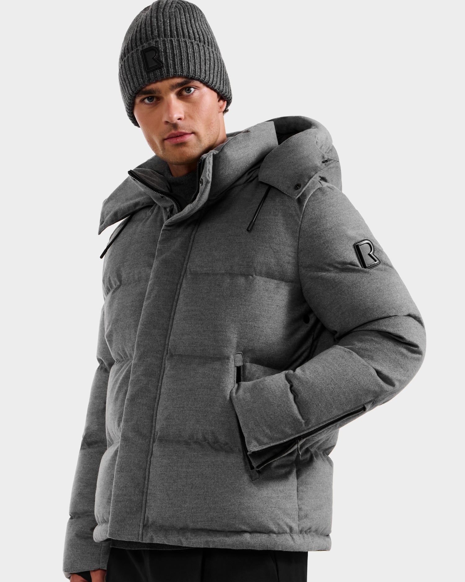 A man in the Rudsak JOEL W - 6125683 Heather Charcoal wool-blend down puffer with removable hood and matching gray knit beanie, both with a black R logo, stands against a plain light gray background.