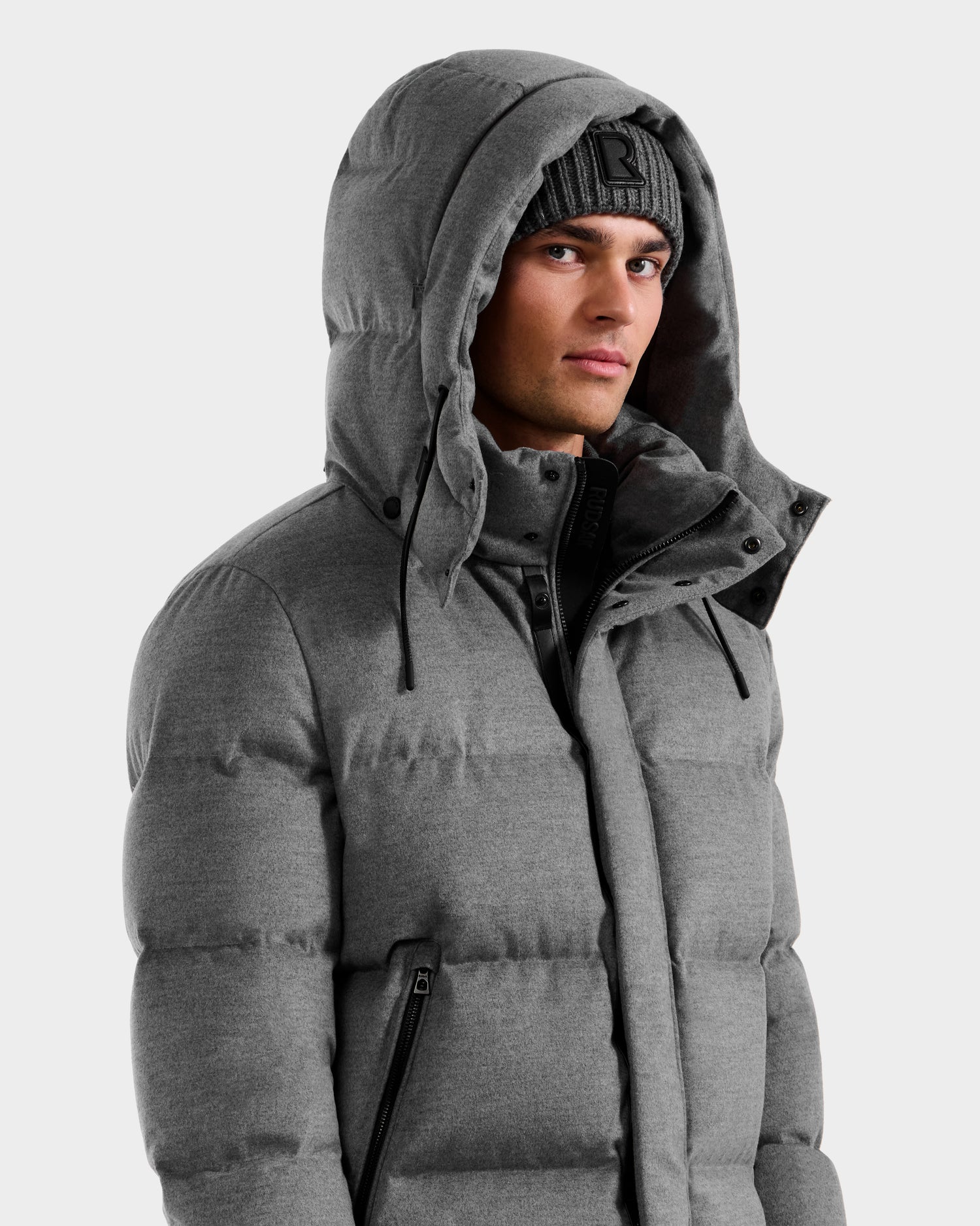 A man wears the Rudsak JOEL W - 6125683 Heather Charcoal wool-blend down puffer with the hood up and a gray beanie, standing against a plain light background and looking slightly to the side with a neutral expression.