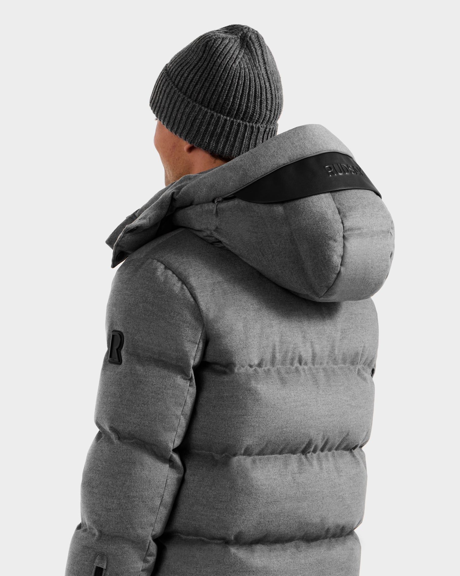 A person wearing the Rudsak JOEL W - 6125683 Heather Charcoal men’s wool-blend down puffer jacket with removable hood and a gray knit beanie, shown from the back and side, against a plain light background.