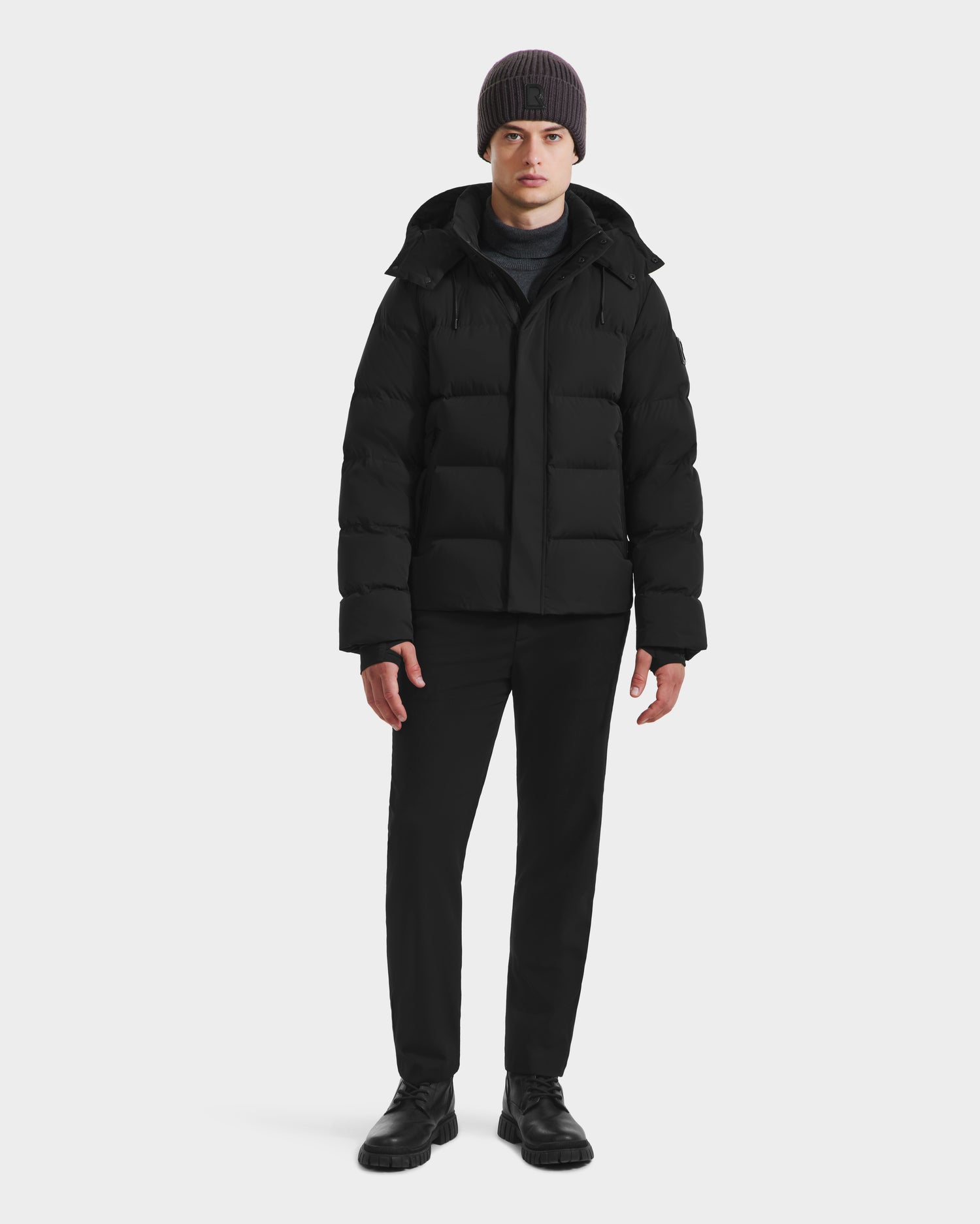 A man faces forward against a white background wearing the Rudsak JOEL - 6124605 Black men’s puffer jacket, black pants, black shoes, and a gray knit beanie, ready for cold weather.