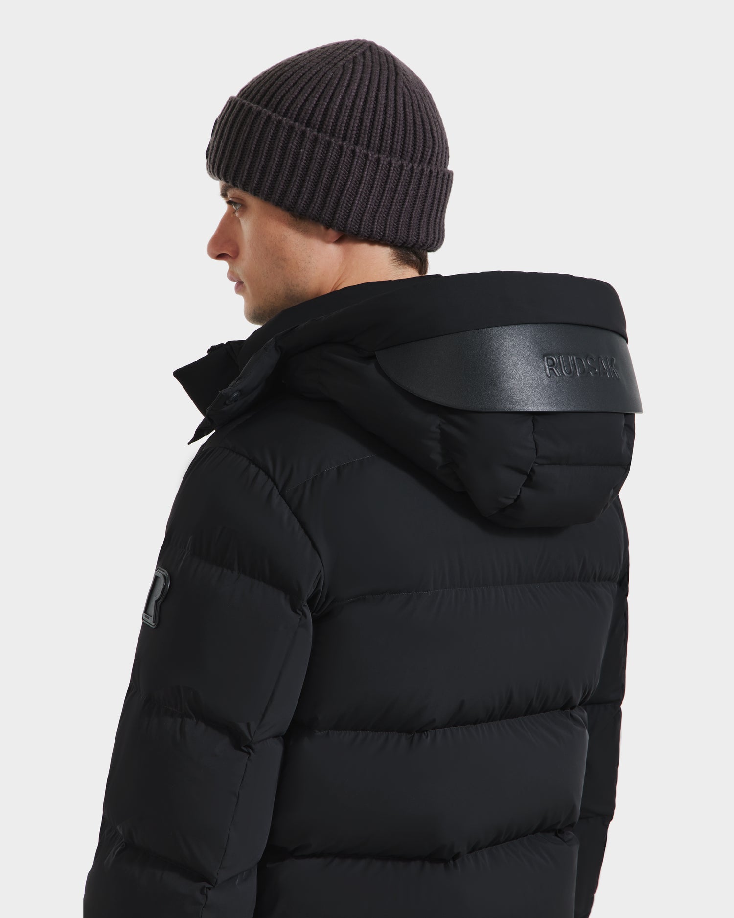 A man faces away in the Rudsak JOEL - 6124605 Black puffer jacket with a hood and a ribbed knit beanie, set against a white background—designed for superior cold-weather performance.