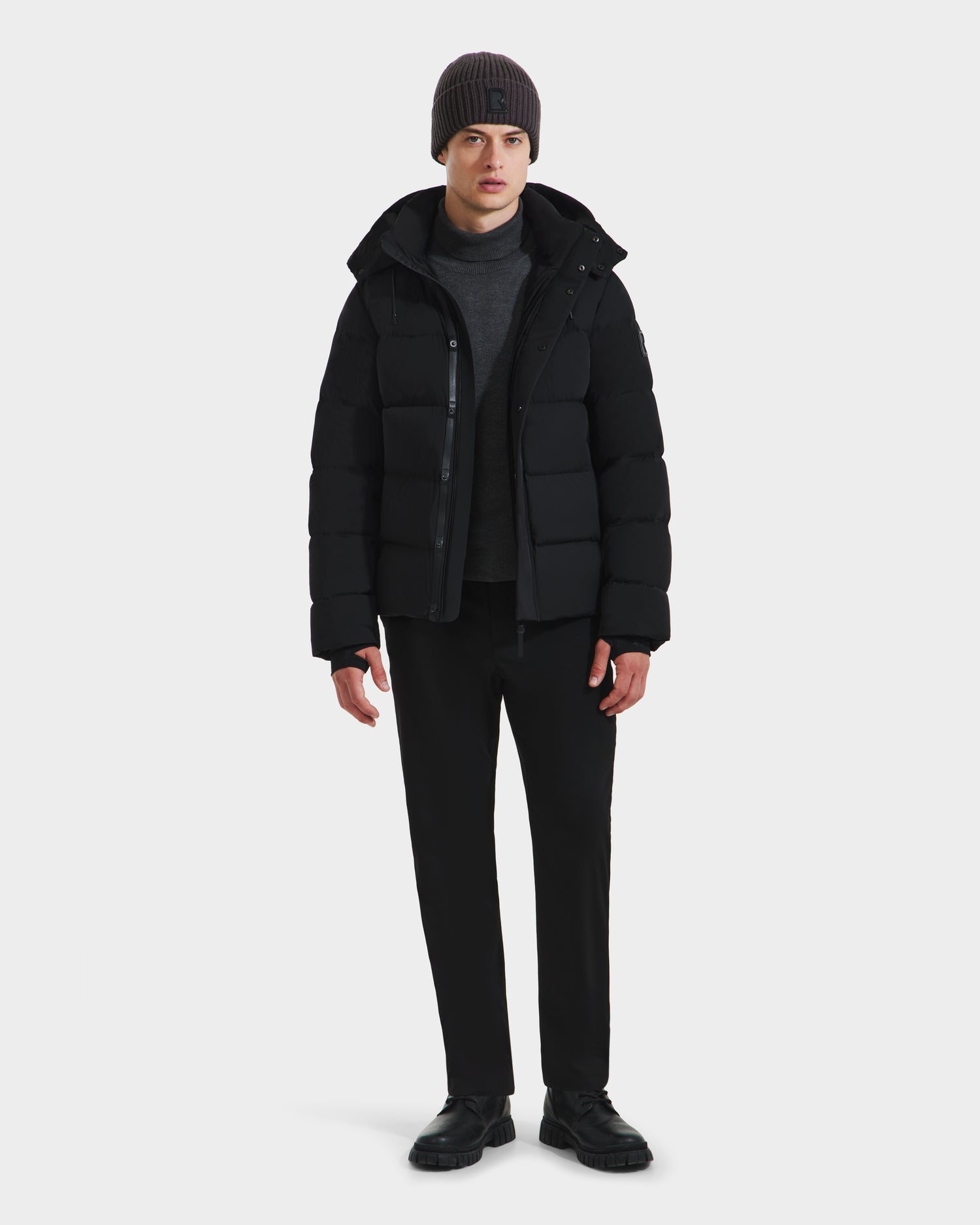 A person stands facing forward wearing the Rudsak JOEL - 6124605 Black men’s puffer jacket, gray beanie, charcoal sweater, black pants, and black shoes against a white background—showcasing cold-weather style.