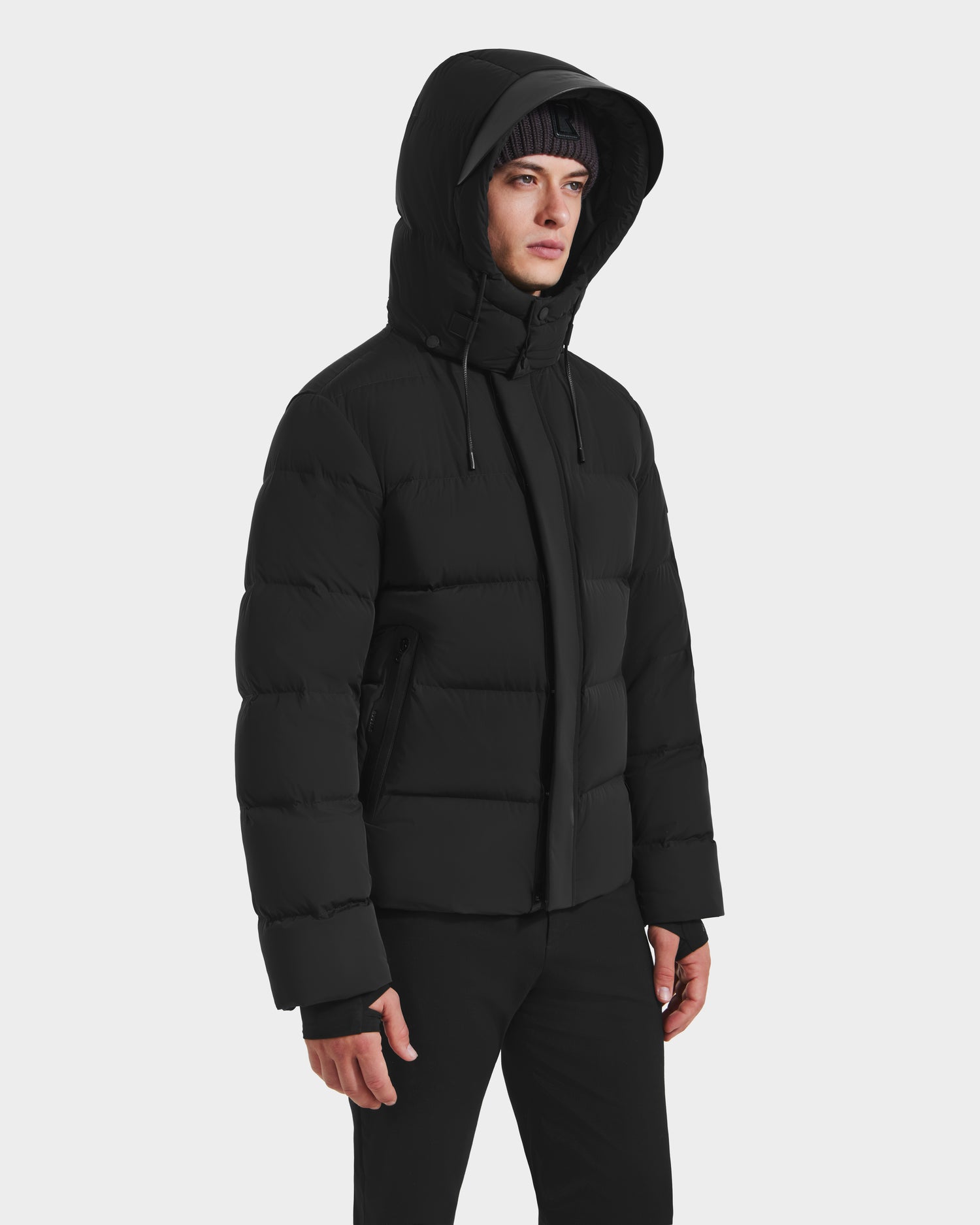 A person wears the Rudsak JOEL - 6124605 Black men’s puffer jacket with a hood and black pants, standing against a white background and looking to the side, highlighting stylish cold-weather performance.
