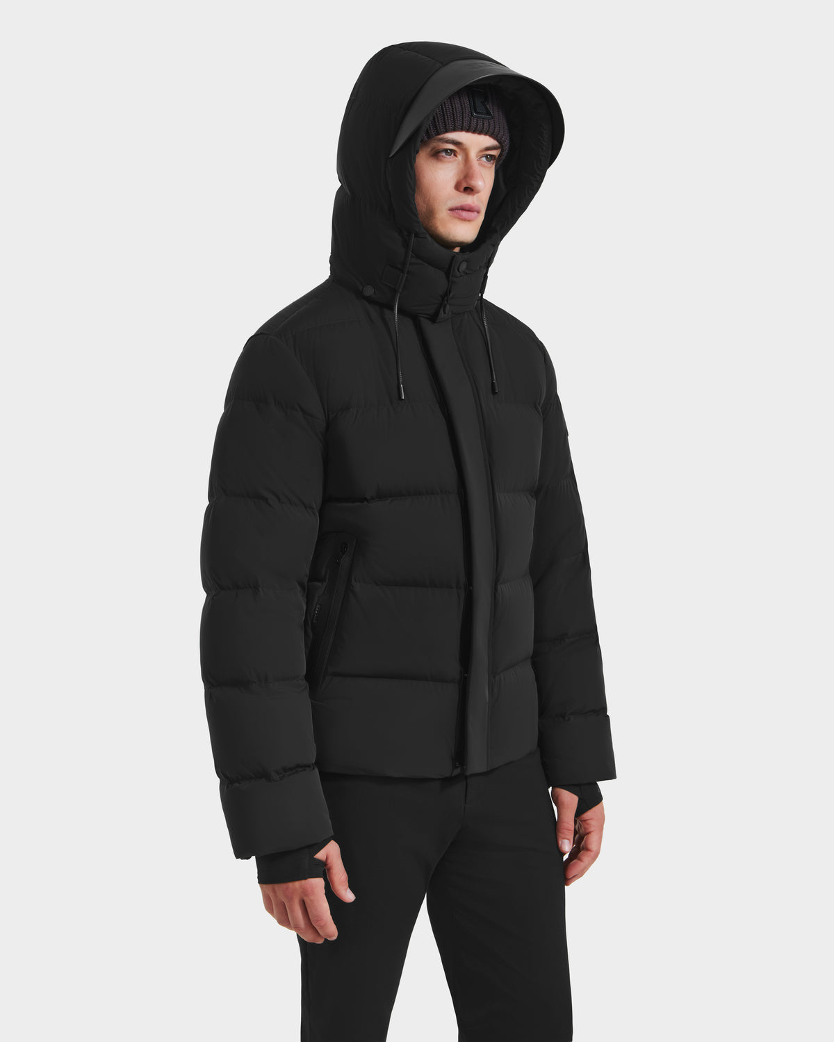 A person wears the Rudsak JOEL - 6124605 Black men’s puffer jacket with a hood and black pants, standing against a white background and looking to the side, highlighting stylish cold-weather performance.