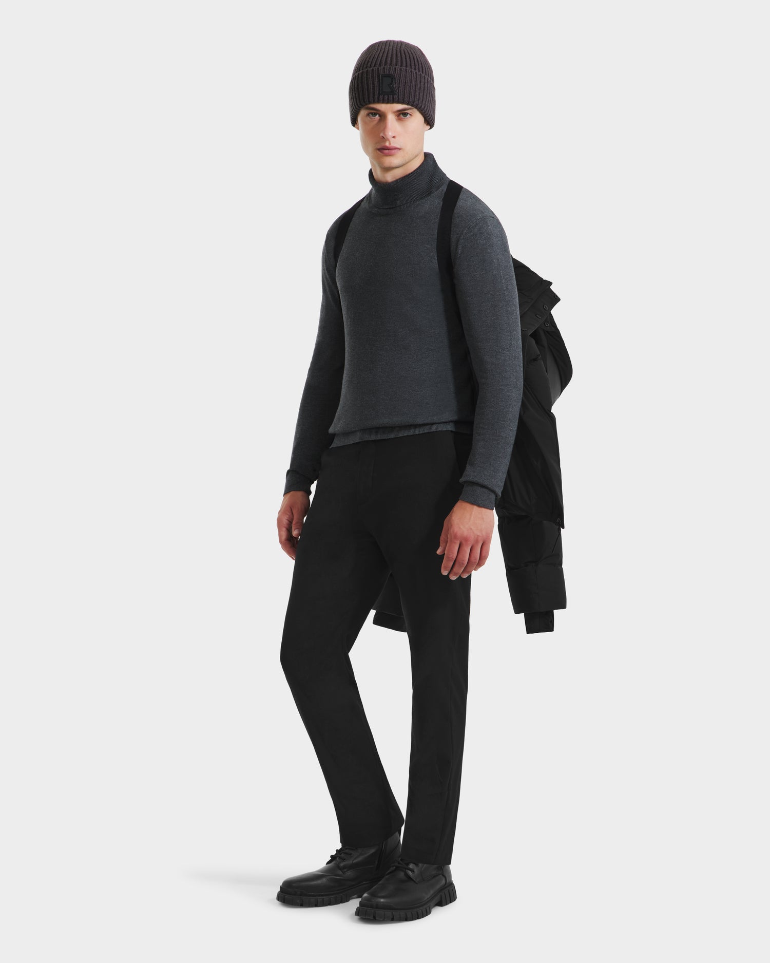 A man stands facing forward against a white background, holding the Rudsak JOEL - 6124605 Black men’s puffer jacket over his shoulder, dressed for cold weather in a gray turtleneck, dark pants, black shoes, and a dark beanie.