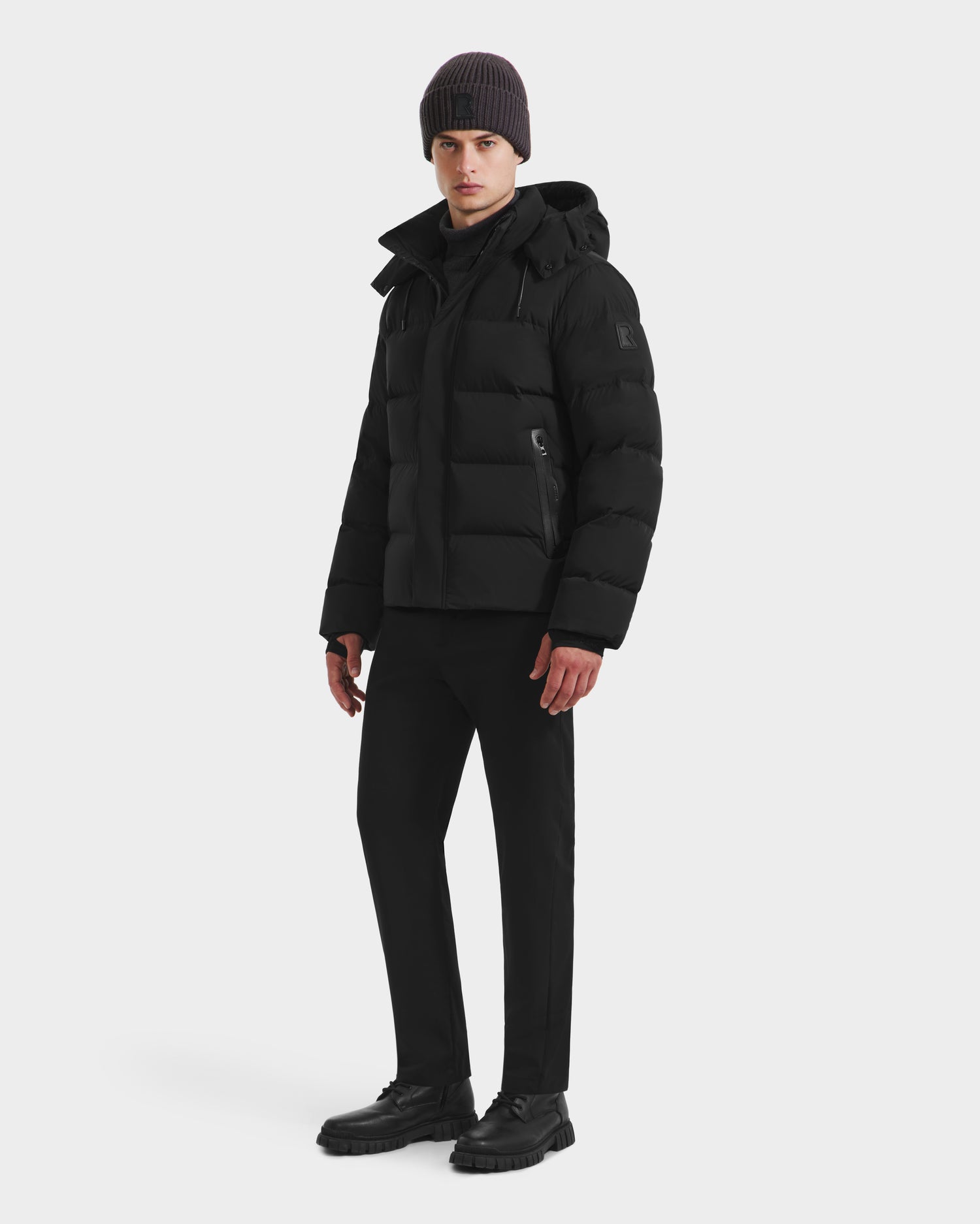 A man wearing the Rudsak JOEL - 6124605 Black men’s puffer jacket, black pants, black shoes, and a dark knit beanie stands angled against a plain white background, highlighting cold-weather performance style.