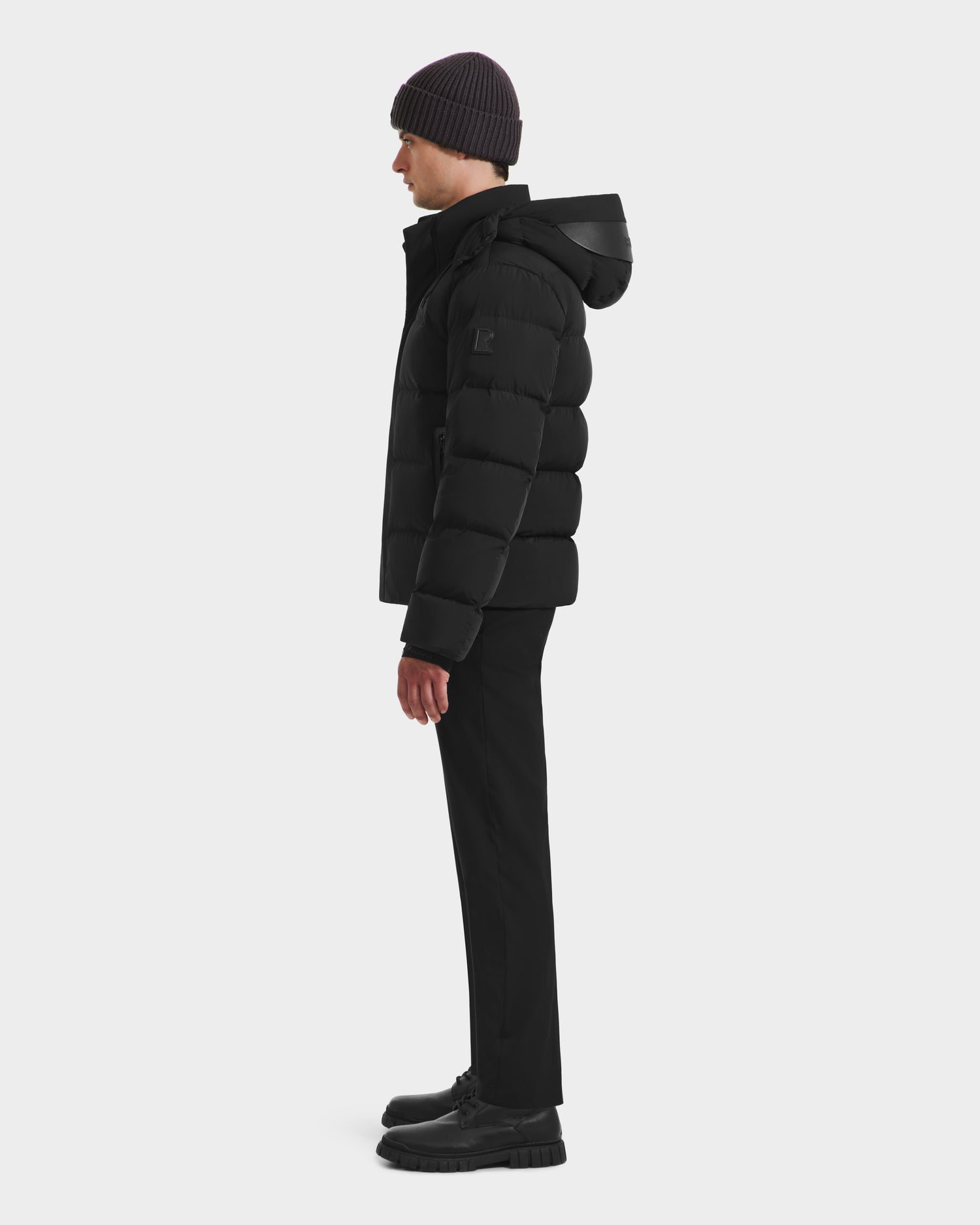 A man stands in profile against a plain white background, wearing the Rudsak JOEL - 6124605 Black mens puffer jacket, paired with a black beanie, trousers, and shoes.