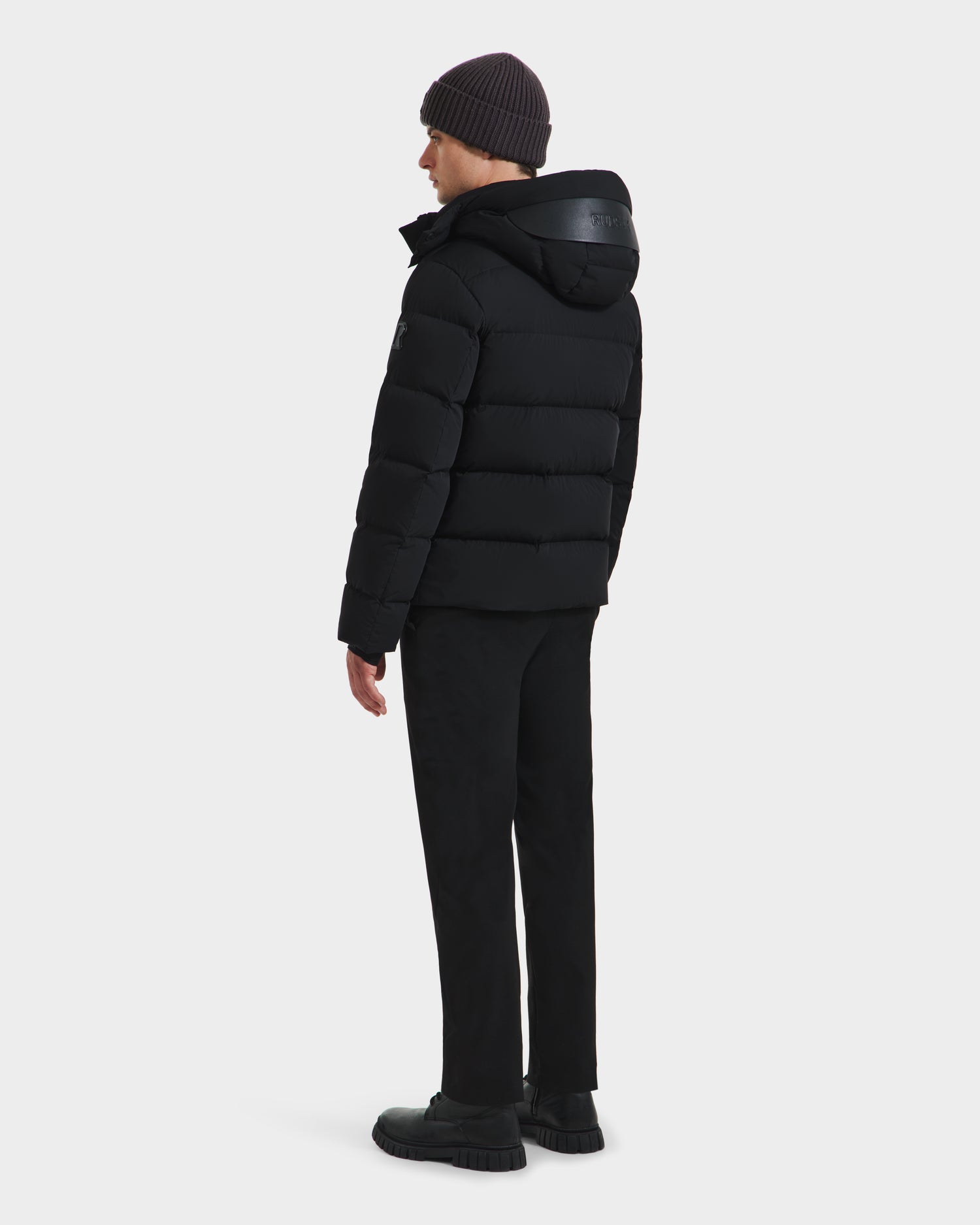 A person faces away, wearing the Rudsak JOEL - 6124605 Black men’s puffer jacket with hood, paired with black pants, shoes, and a dark knit beanie—ideal for cold weather—set against a plain white background.