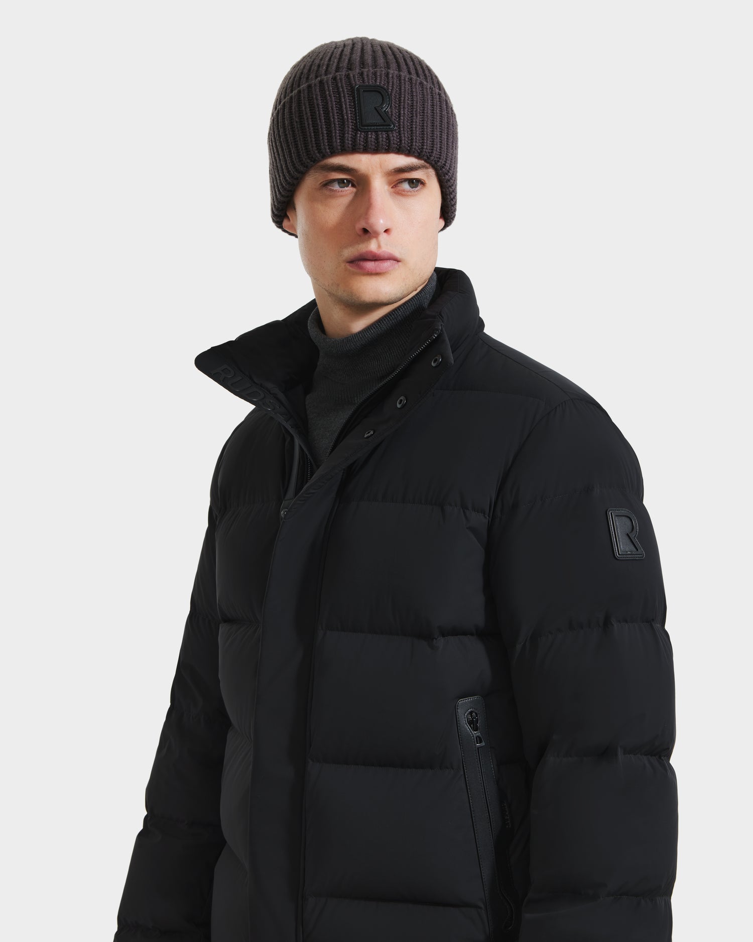 A young man in Rudsak’s JOEL - 6124605 Black men’s puffer jacket and a ribbed knit beanie, both featuring a subtle R logo, looks slightly to the side against a plain light background.