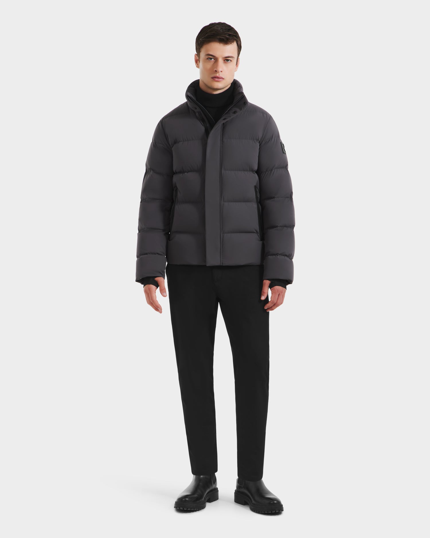 A young man faces forward, wearing the Rudsak JOEL - 6124605 Grey men's puffer jacket, paired with a black turtleneck, pants, and shoes against a plain white background.