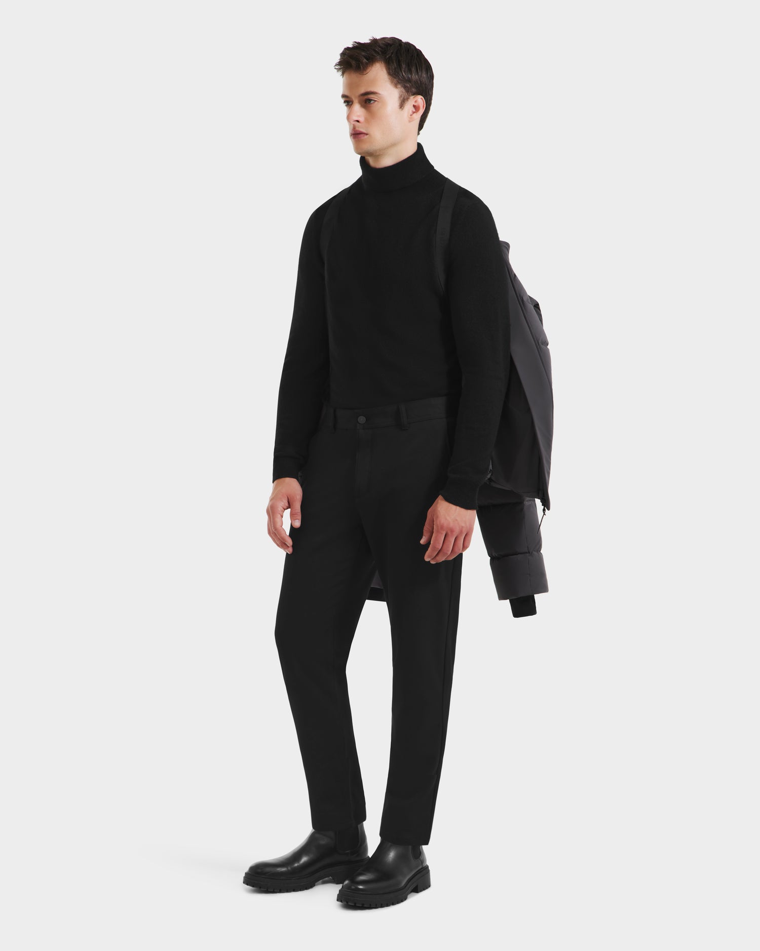 A man poses against a white background wearing a black turtleneck, pants, and boots, with the Rudsak JOEL - 6124605 Grey down-filled men’s puffer jacket draped over his shoulder.