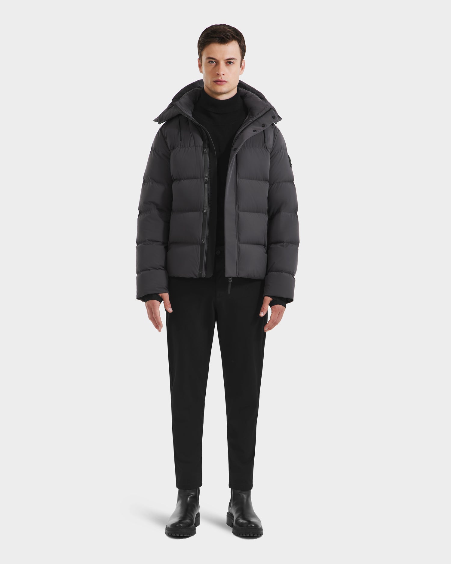 A man stands facing forward, wearing the Rudsak JOEL - 6124605 Grey men’s puffer jacket over a black turtleneck, black pants, and black boots against a plain light background.