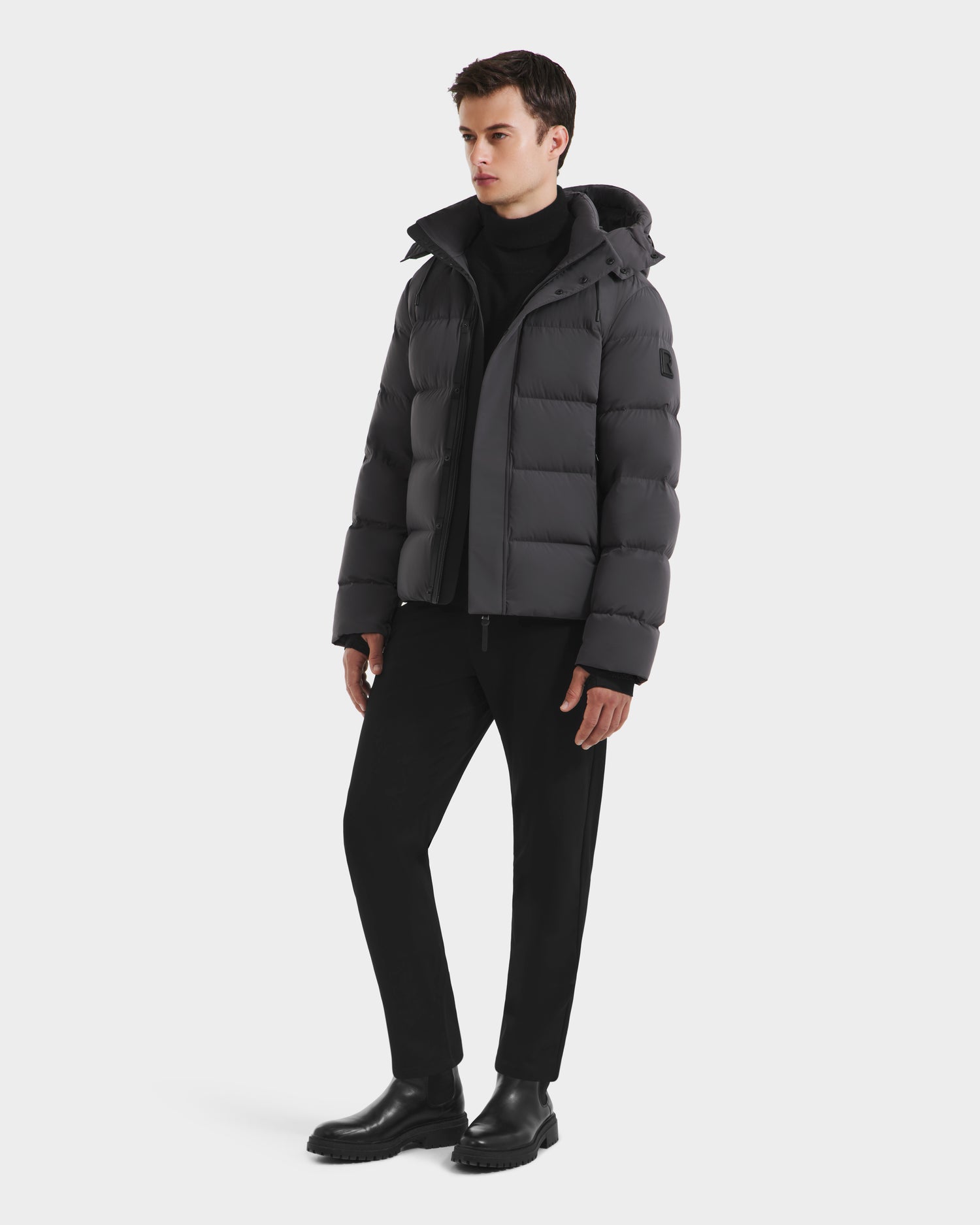 A young man stands against a white background wearing the Rudsak JOEL - 6124605 Grey down-filled men’s puffer jacket, styled with a black turtleneck, pants, and boots. He faces slightly to the side with a neutral expression, ready for cold weather.