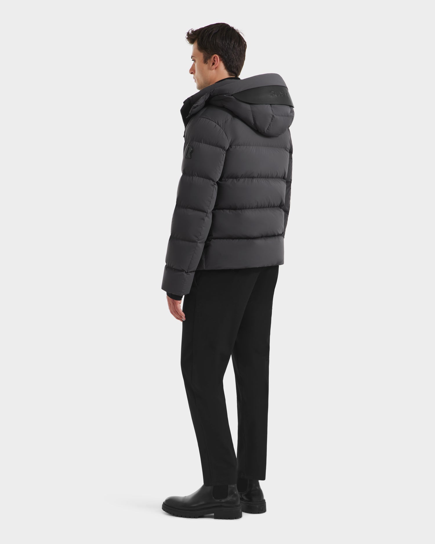 A man faces away, wearing the Rudsak JOEL - 6124605 Grey down-filled men’s puffer jacket with a hood, paired with black pants and boots, against a plain white background.