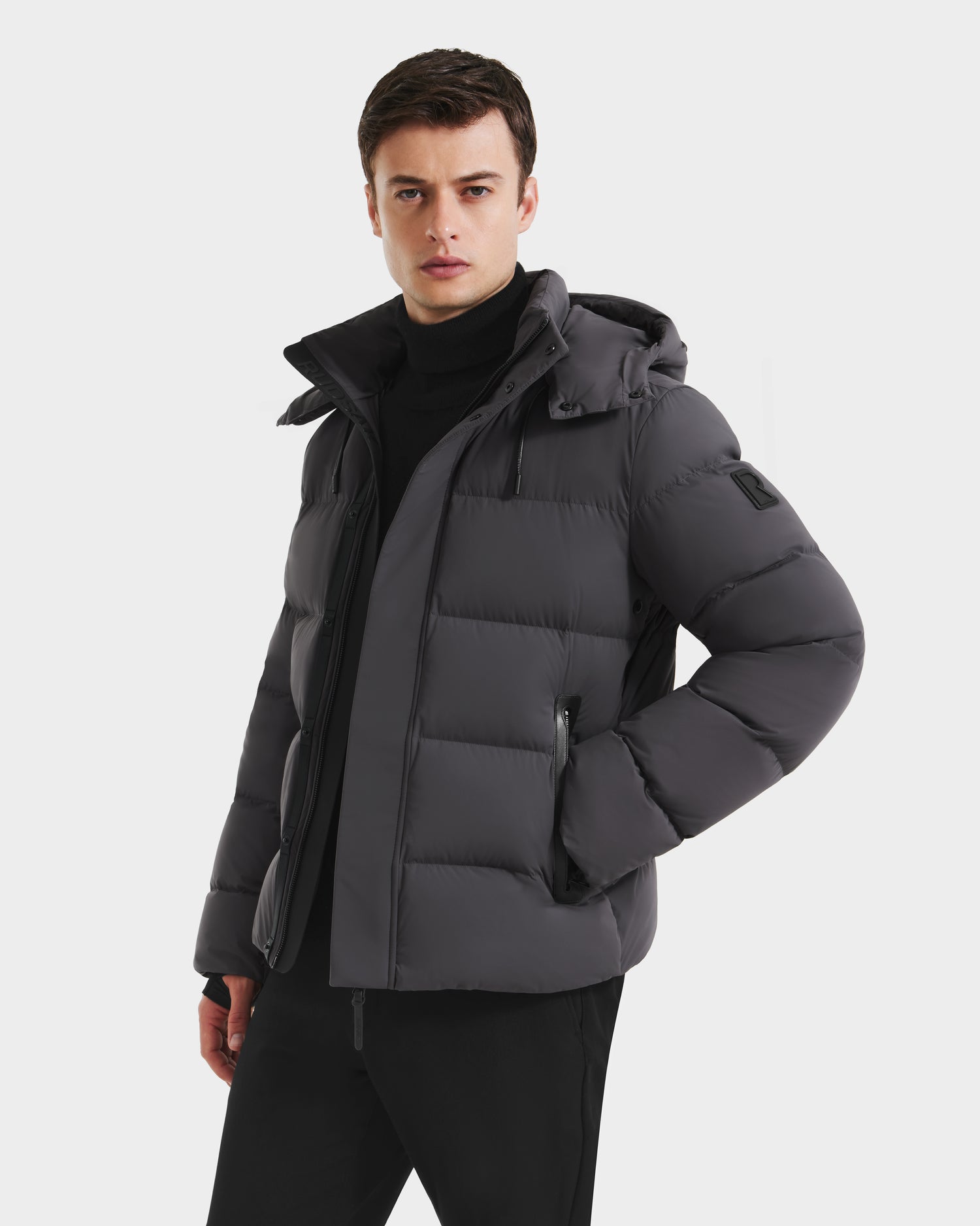 A young man confidently poses against a white background, wearing the Rudsak JOEL - 6124605 Grey men's puffer down jacket with hood and black pants—perfect for cold-weather days.