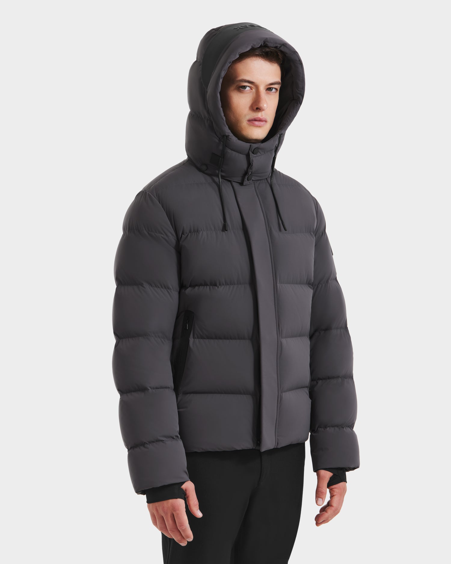 A man wearing the Rudsak JOEL - 6124605 Grey men’s puffer jacket with a hood, designed for cold weather, stands against a plain white background, looking forward with a neutral expression.