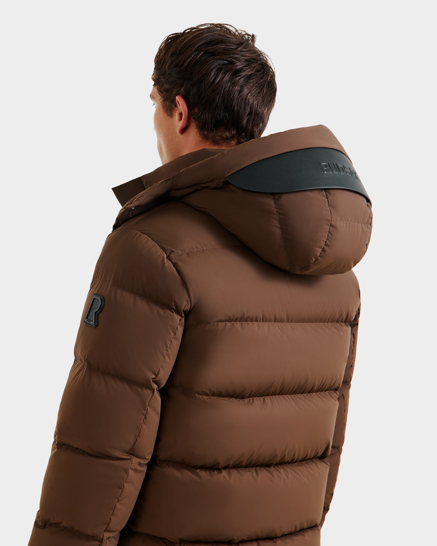 A person is shown from behind wearing the Rudsak JOEL - 6124605 Mocha men’s puffer jacket with hood, featuring a black shoulder patch and black hood accent, set against a plain, light-colored background.