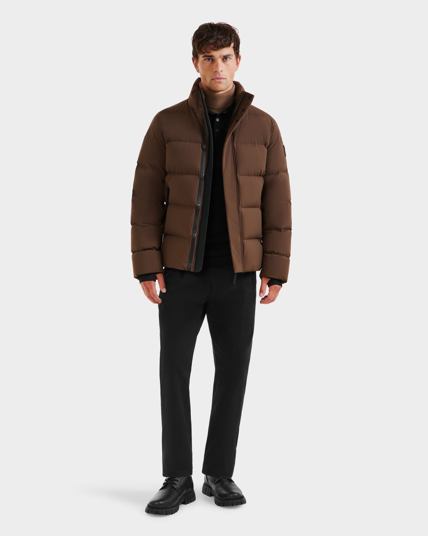 A man stands facing forward, wearing the Rudsak JOEL - 6124605 Mocha weather-resistant men’s puffer jacket over a black shirt and pants with black shoes, against a plain light gray background.