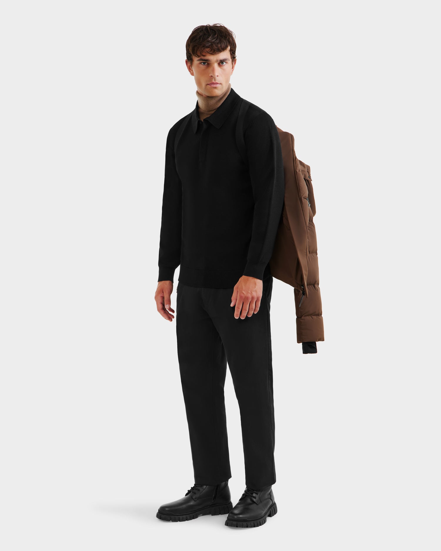A man stands against a plain background wearing a black outfit, with the Rudsak JOEL - 6124605 Mocha weather-resistant men’s puffer jacket draped over one shoulder, looking slightly to his left.