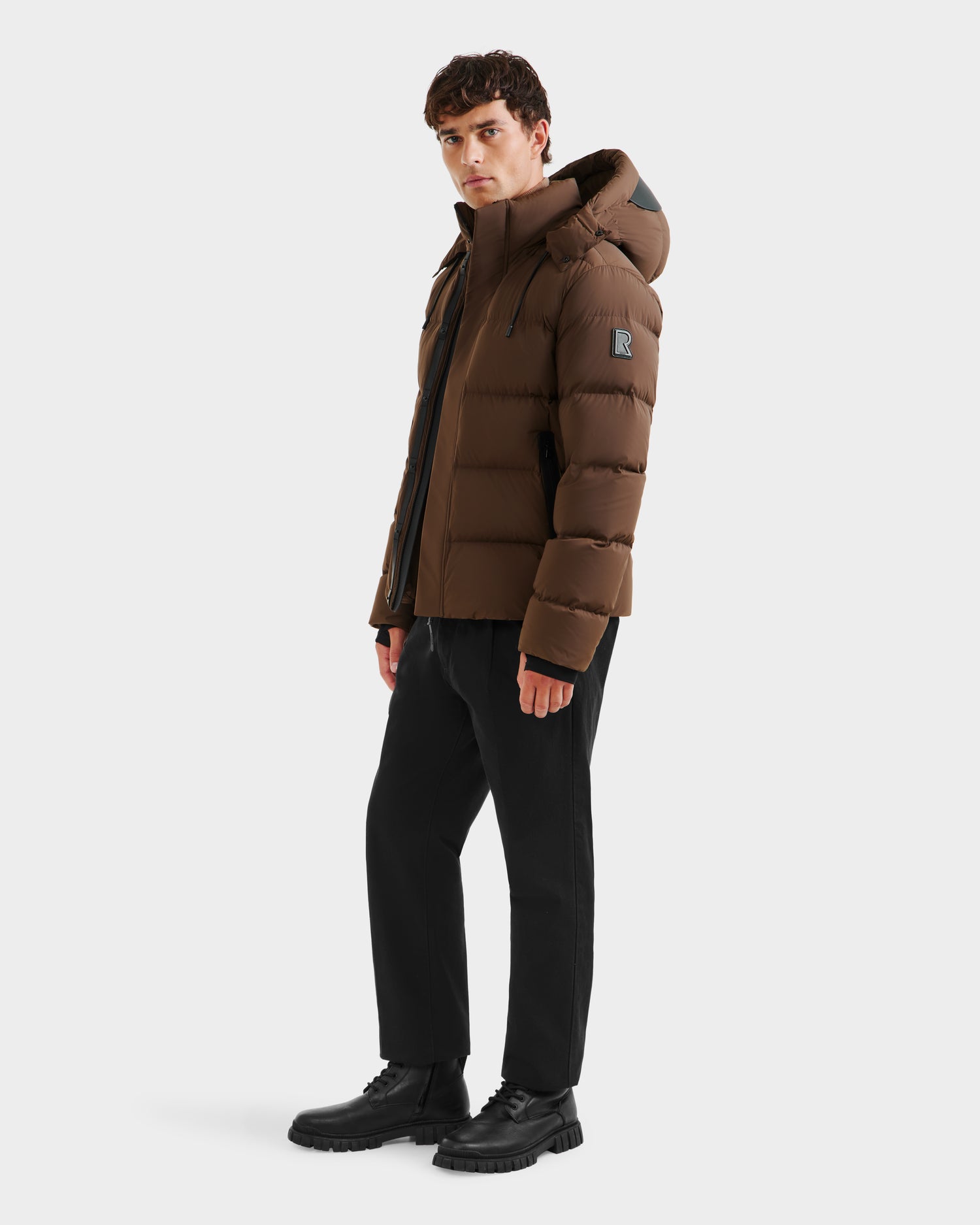A man stands slightly turned, wearing the Rudsak JOEL - 6124605 Mocha weather-resistant men’s puffer jacket with a hood, paired with black pants and boots against a plain white background.