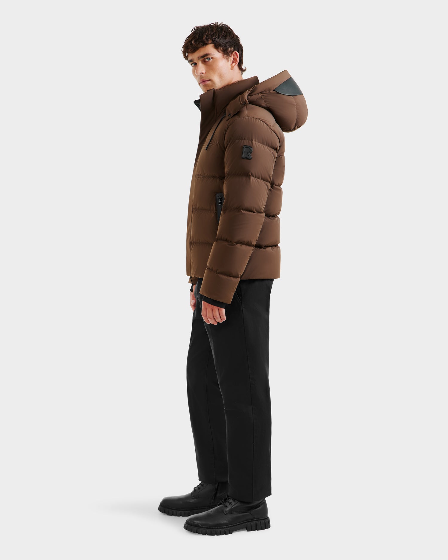 A person stands sideways against a plain light background, wearing the Rudsak JOEL - 6124605 Mocha men’s puffer jacket with a hood, paired with black pants and black shoes.