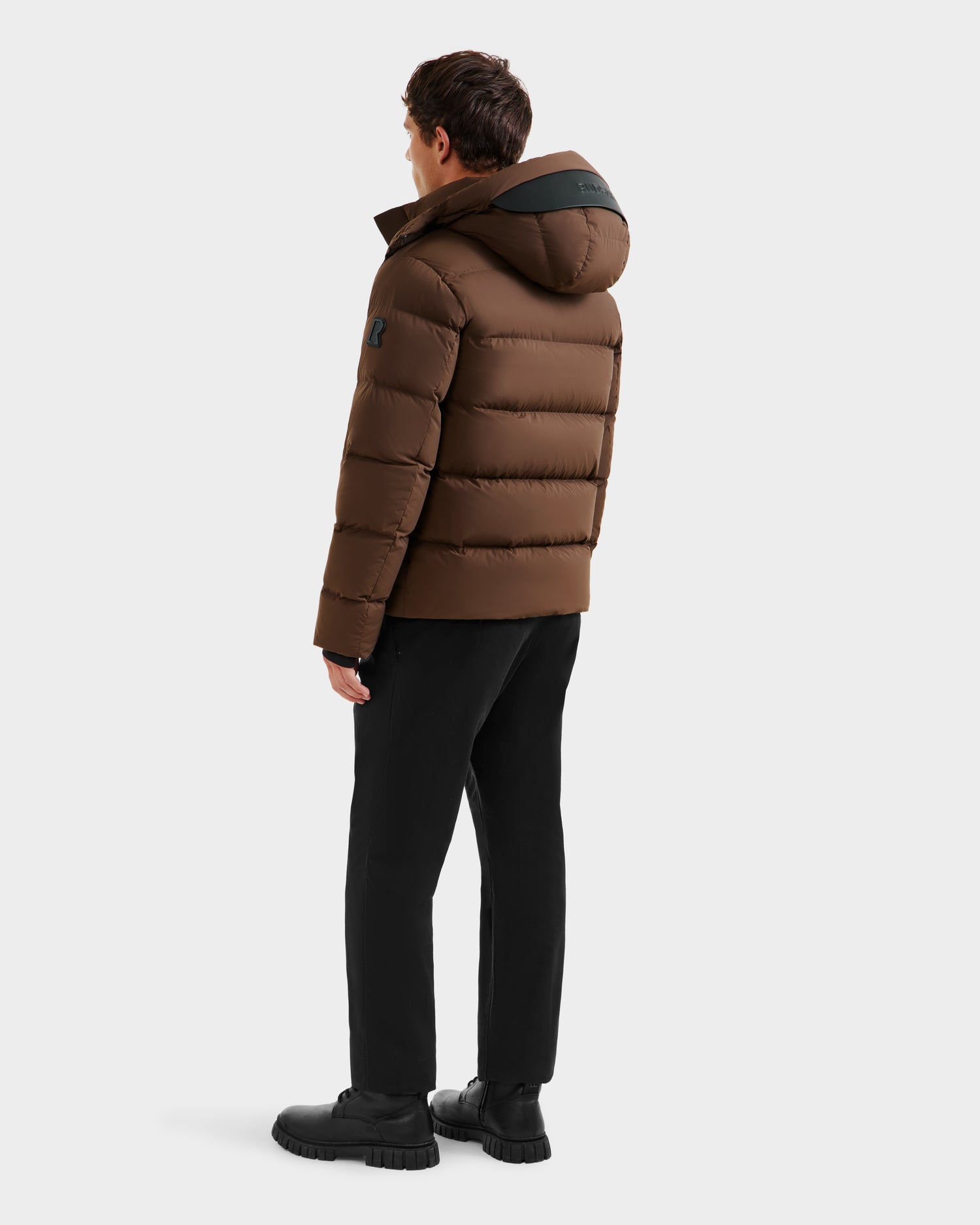 A man stands facing away, wearing the Rudsak JOEL - 6124605 Mocha weather-resistant men’s puffer jacket with hood, black pants, and chunky-soled shoes against a plain white background.