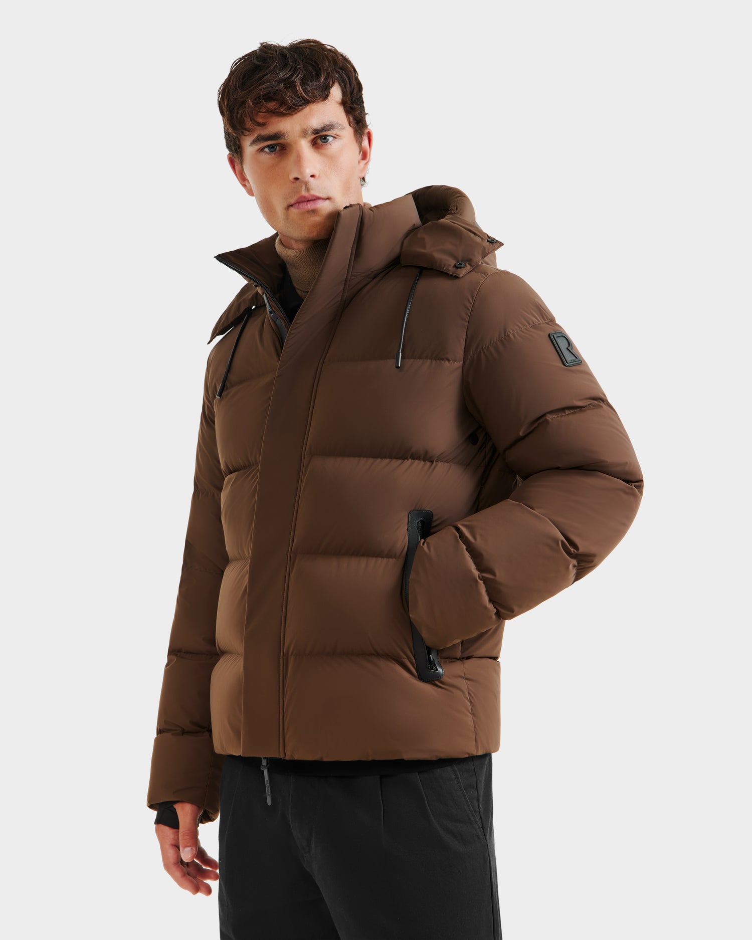 A man stands against a white background, wearing the Rudsak JOEL - 6124605 Mocha brown down-filled men’s puffer jacket with a hood and black zippers, one hand in his pocket, prepared for cold weather.