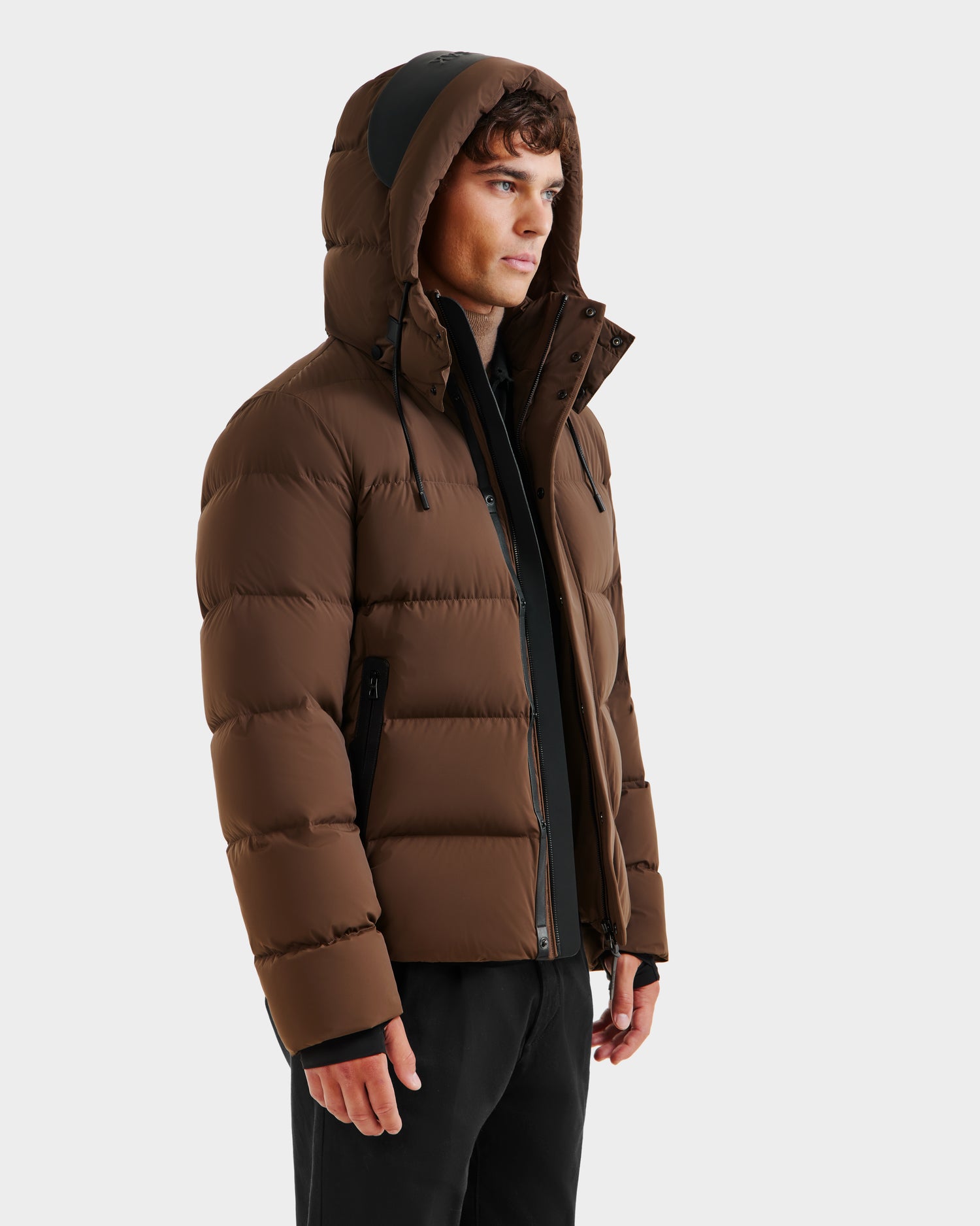 A person with short curly hair wears the Rudsak JOEL - 6124605 Mocha men’s weather-resistant puffer jacket with a hood, zipped up, standing slightly to the side against a plain white background.