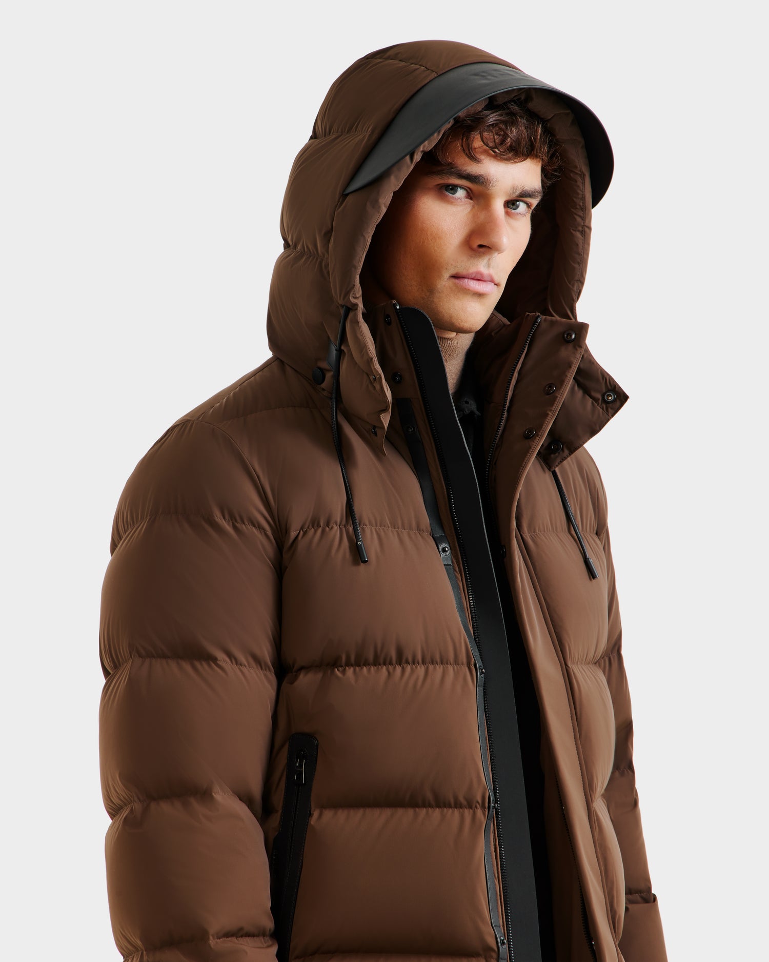 A young man models the Rudsak JOEL - 6124605 Mocha men’s weather-resistant puffer jacket with hood, partially unzipped to show a black inner layer, against a plain light background.