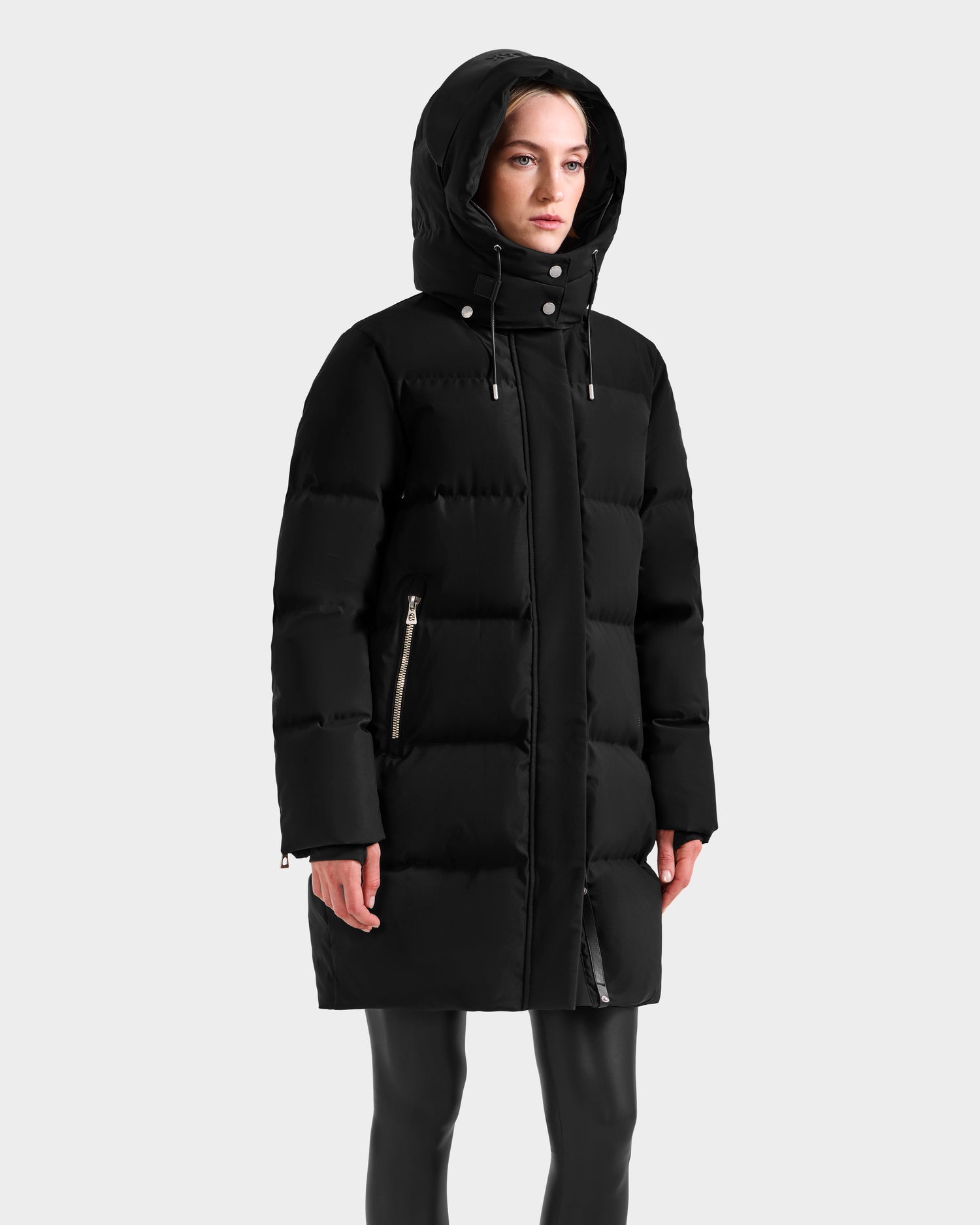 A woman models the Rudsak JOELLE H - 8125602 Black down puffer with a hood and silver zipper pockets, paired with black pants, against a plain light gray background.