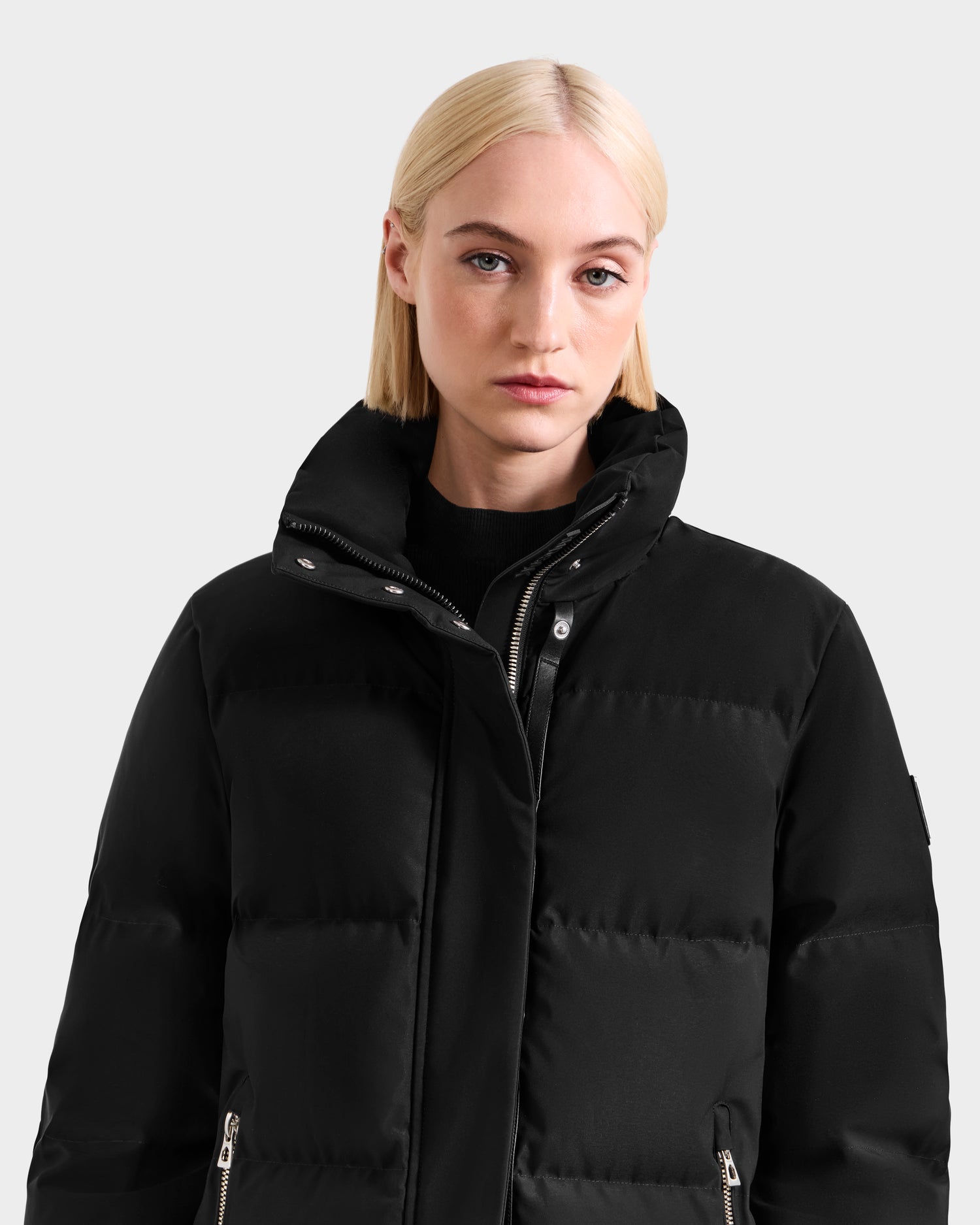 A woman with straight blonde hair wears the Rudsak JOELLE H - 8125602 Black women’s down puffer with a high collar, standing against a light gray background and looking at the camera with a neutral expression.