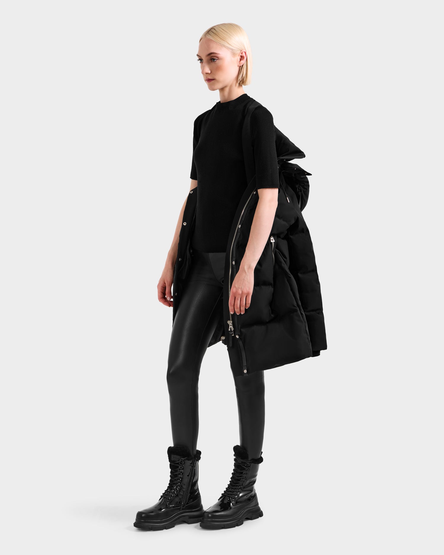 A person with short blond hair wears Rudsak’s JOELLE H - 8125602 Black women’s down puffer over one shoulder, styled with a black top, faux leather pants, and lace-up boots against a plain light gray background.