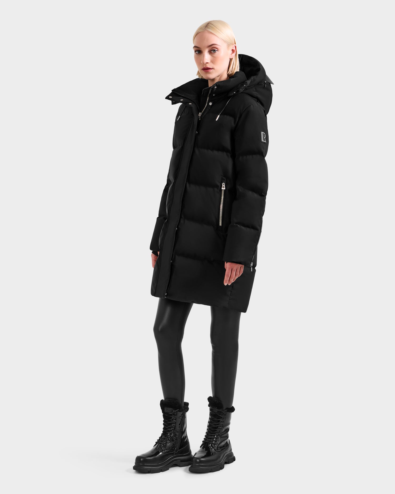 A person with short blonde hair wears the Rudsak JOELLE H - 8125602 Black down puffer with a hood, black leggings, and chunky lace-up boots, standing against a plain light grey background.